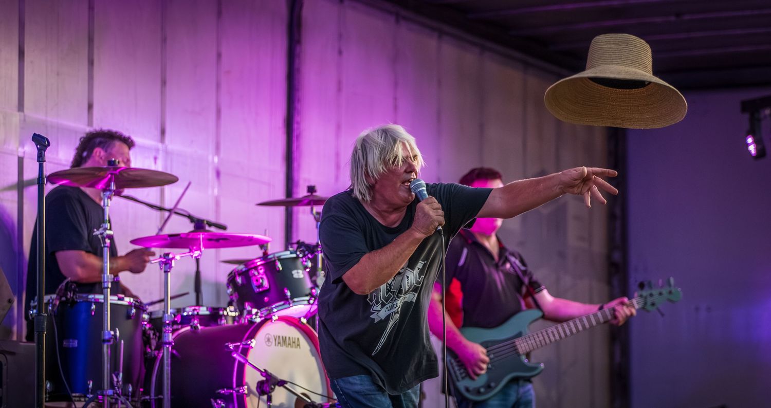 Rock band performs on stage with purple lighting as singer points outward while drummer and bassist play behind them.