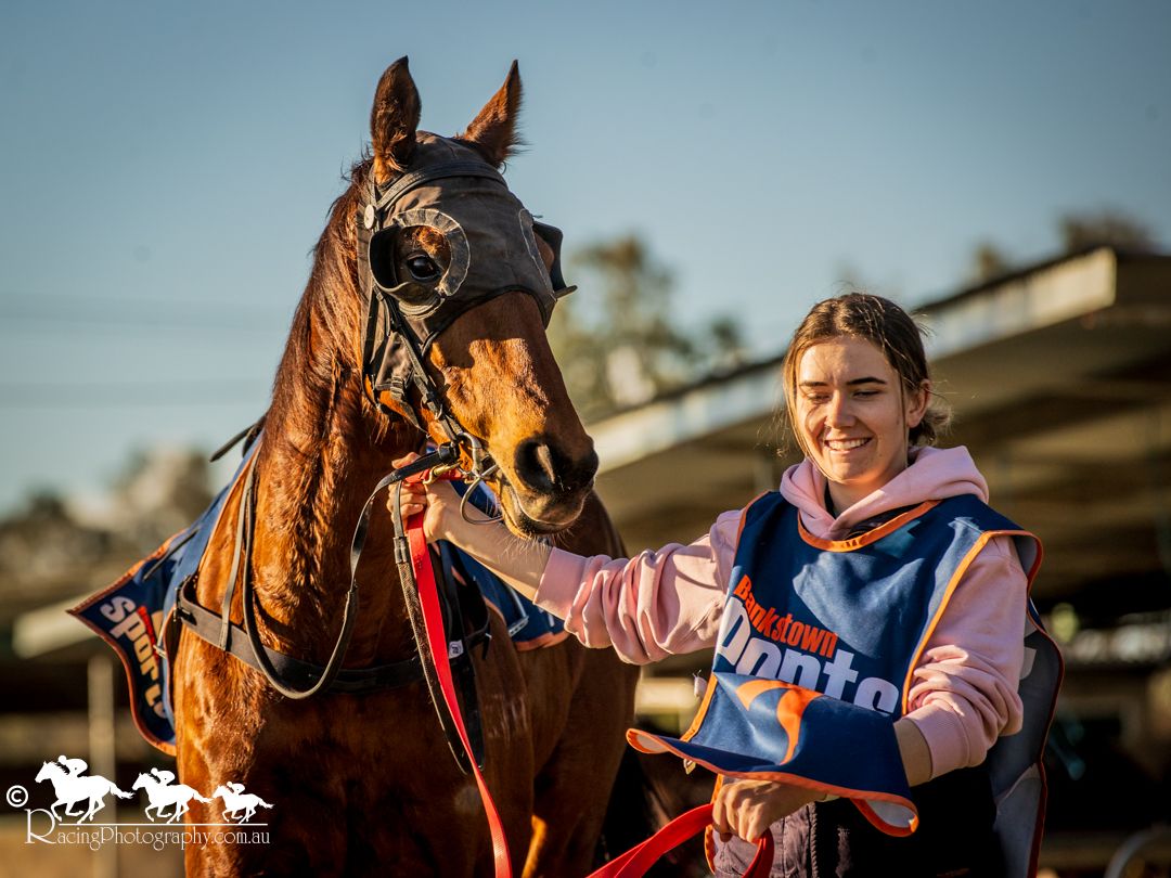 Young woman smiling beside a bay horse at a racetrack, wearing a pink hoodie and holding the horse's lead.