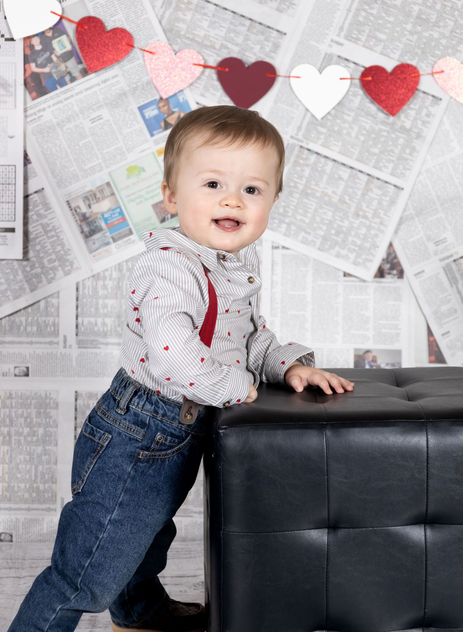 Baby in jeans and white shirt leans against black leather furniture with red heart decorations and newspaper background.