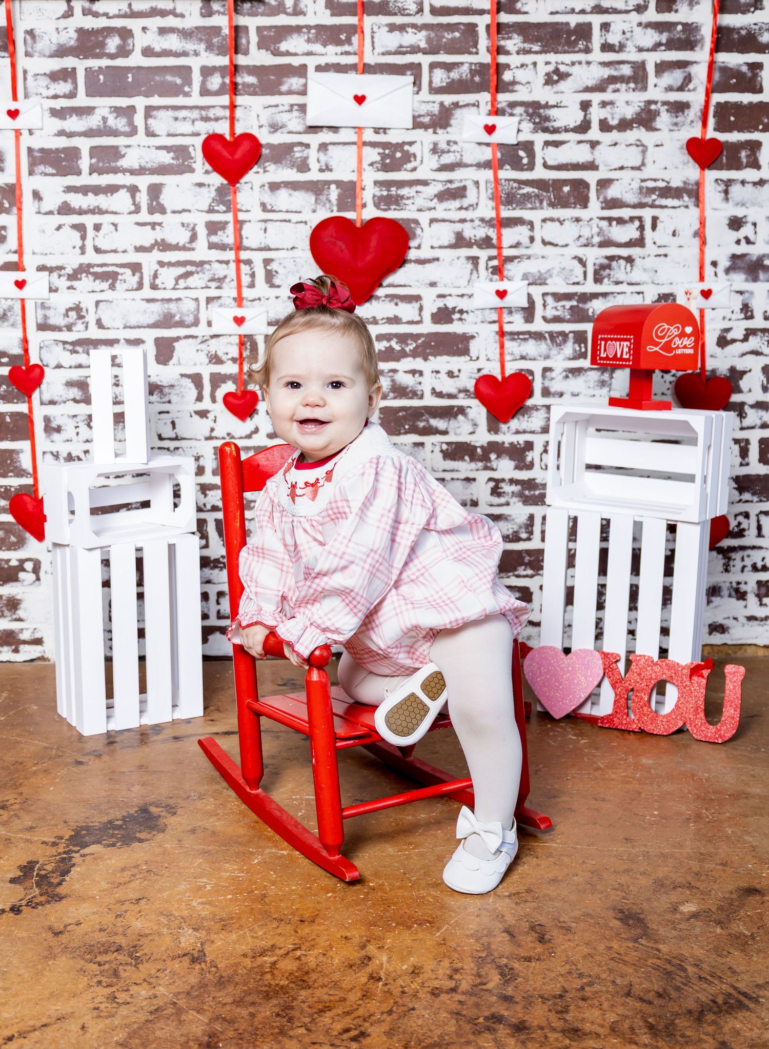 Valentine's Day photo setup with white wooden rocking chairs, red hearts, and rustic brick wall backdrop.