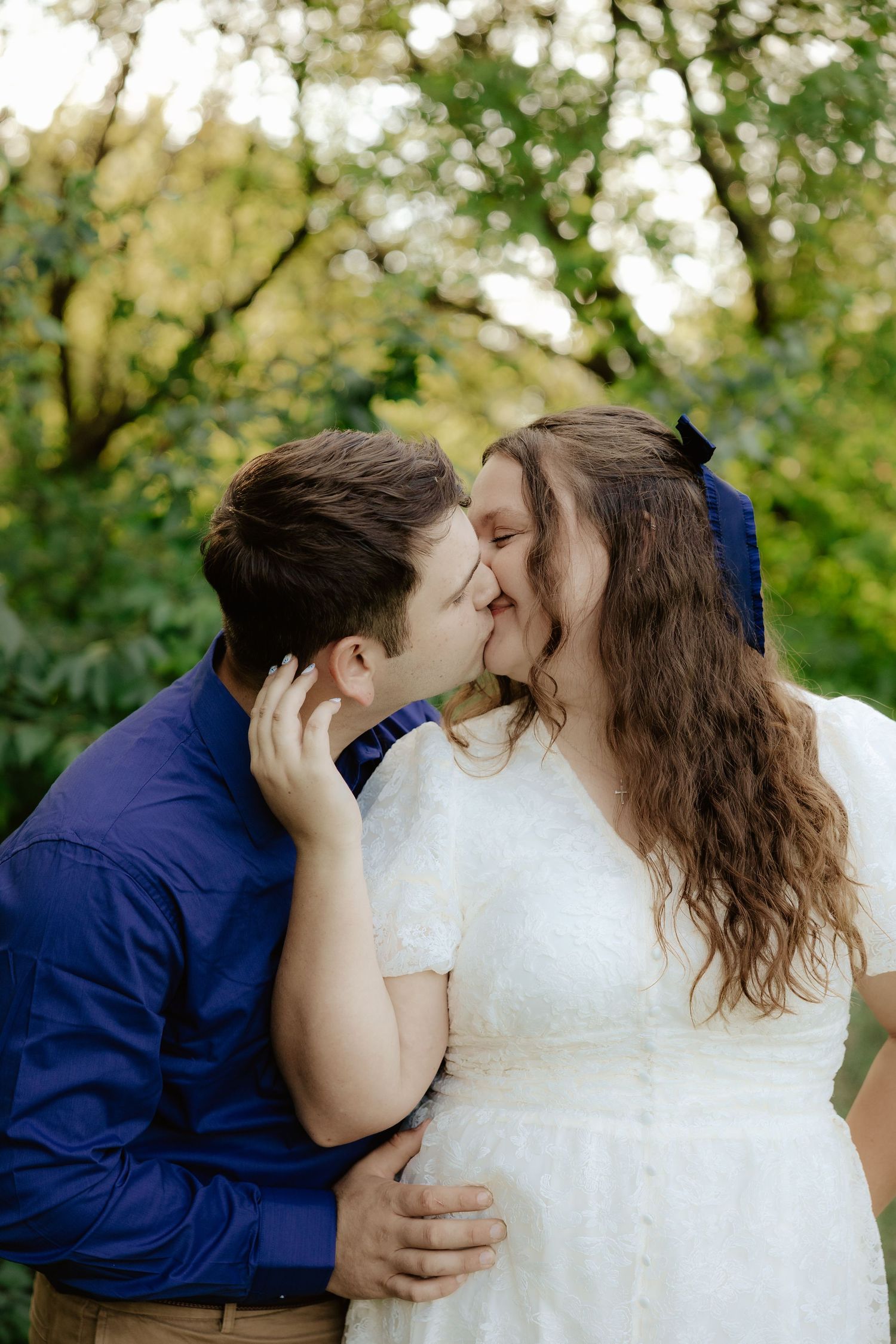 A couple poses together outdoors in wedding attire with white dress and navy blue shirt among green foliage.