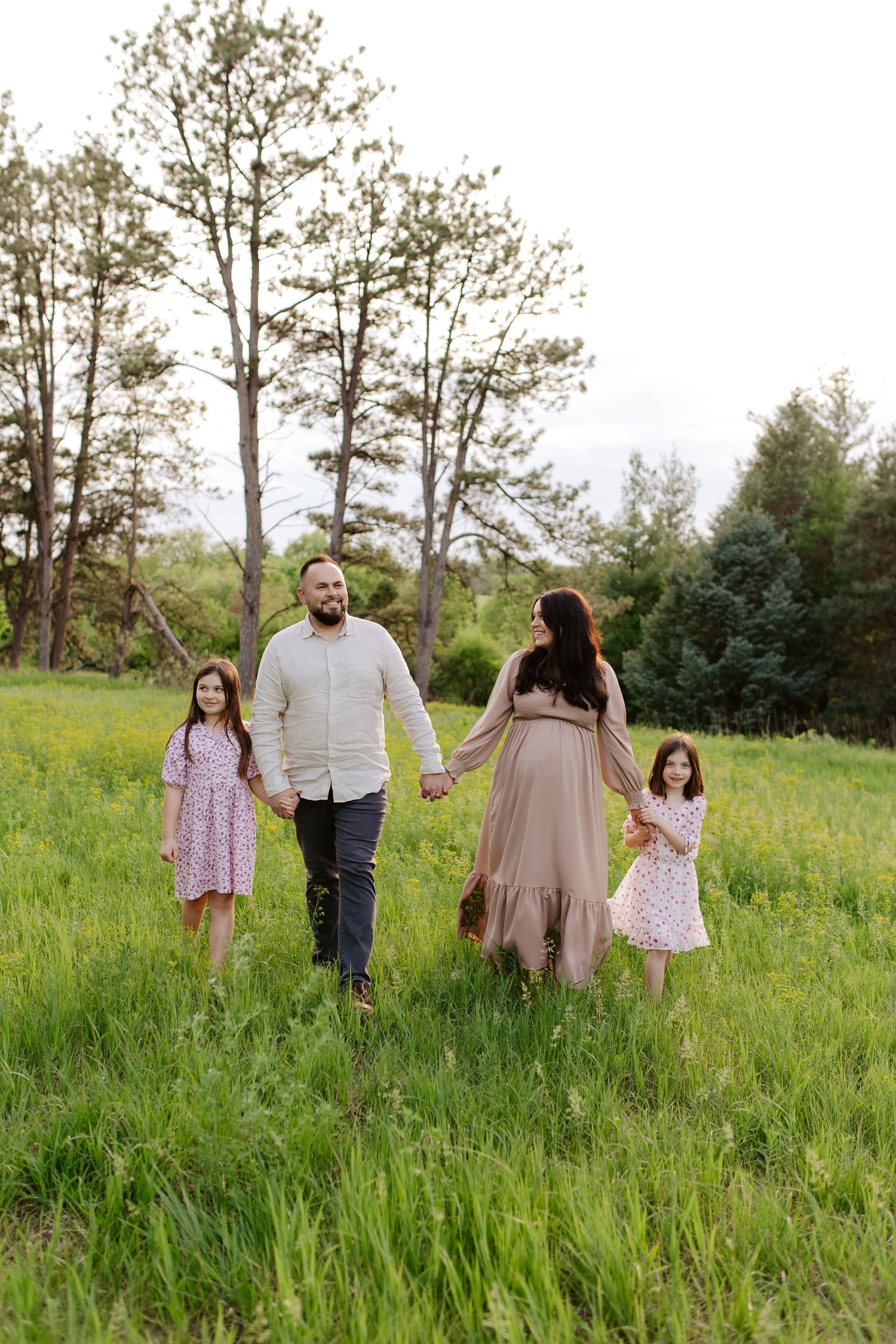 A family walks together holding hands in a green meadow with tall trees in the background on a summer day, maternity photoshoot.