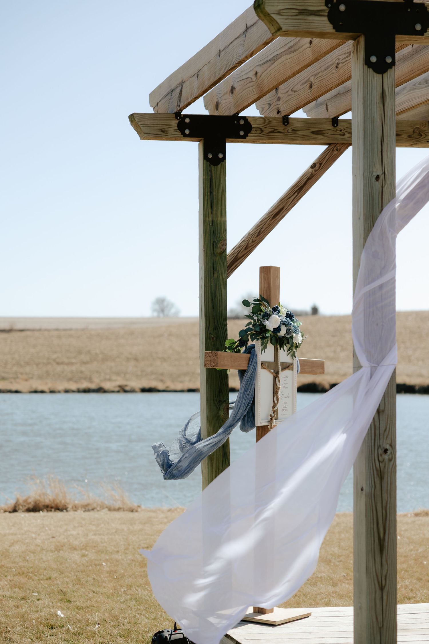 Outdoor wedding altar with wooden cross, white flowing fabric, and blue floral decor beside a serene pond at Sparks Barn in Nebraska