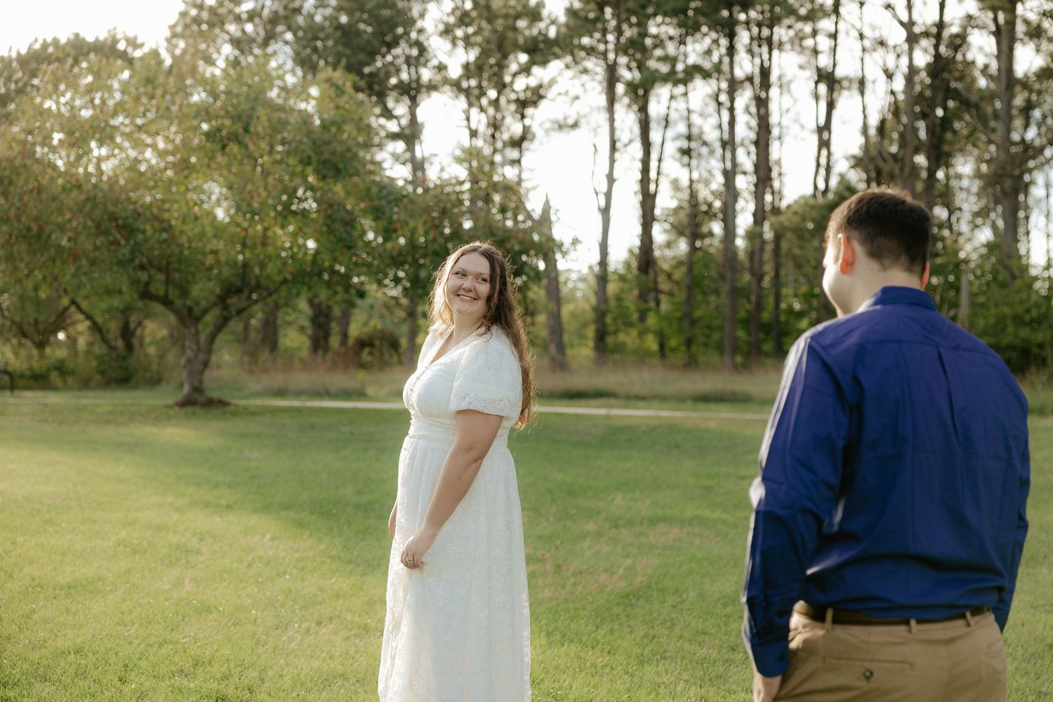 A couple wearing white and navy blue outfits walks together in a grassy field surrounded by pine trees.
