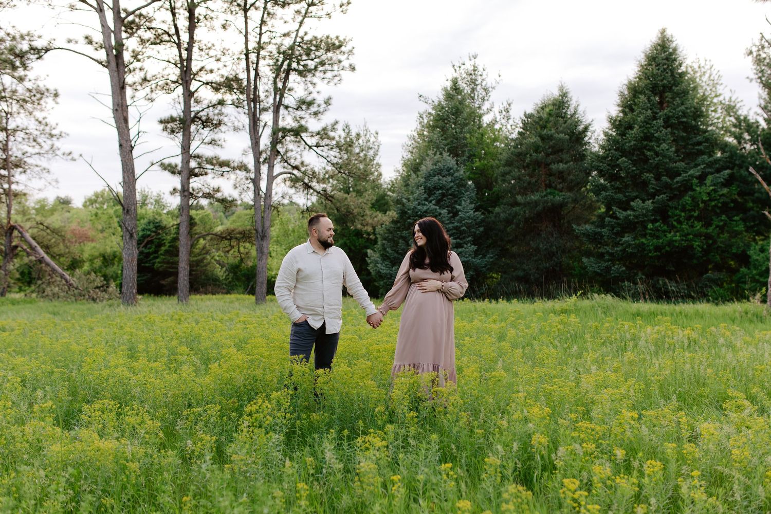 Two people hold hands while standing in a lush meadow of yellow wildflowers with evergreen trees in the background.