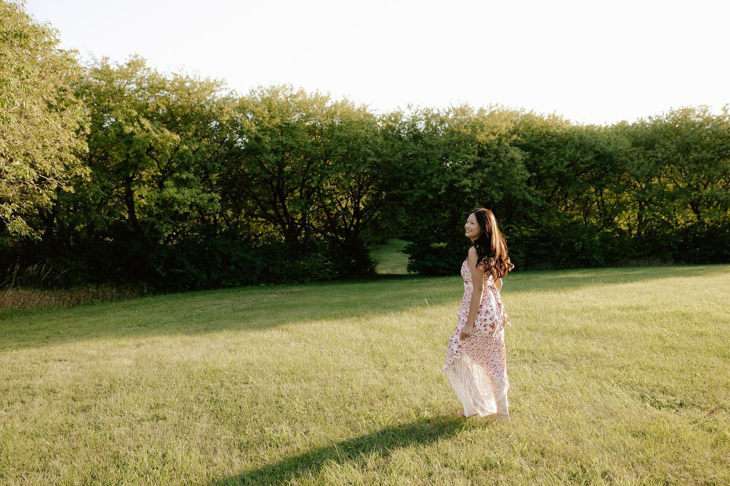 A girl in a white floral dress twirls playfully in a sunlit meadow surrounded by green trees at Pioneers Park in Lincoln Nebraska.