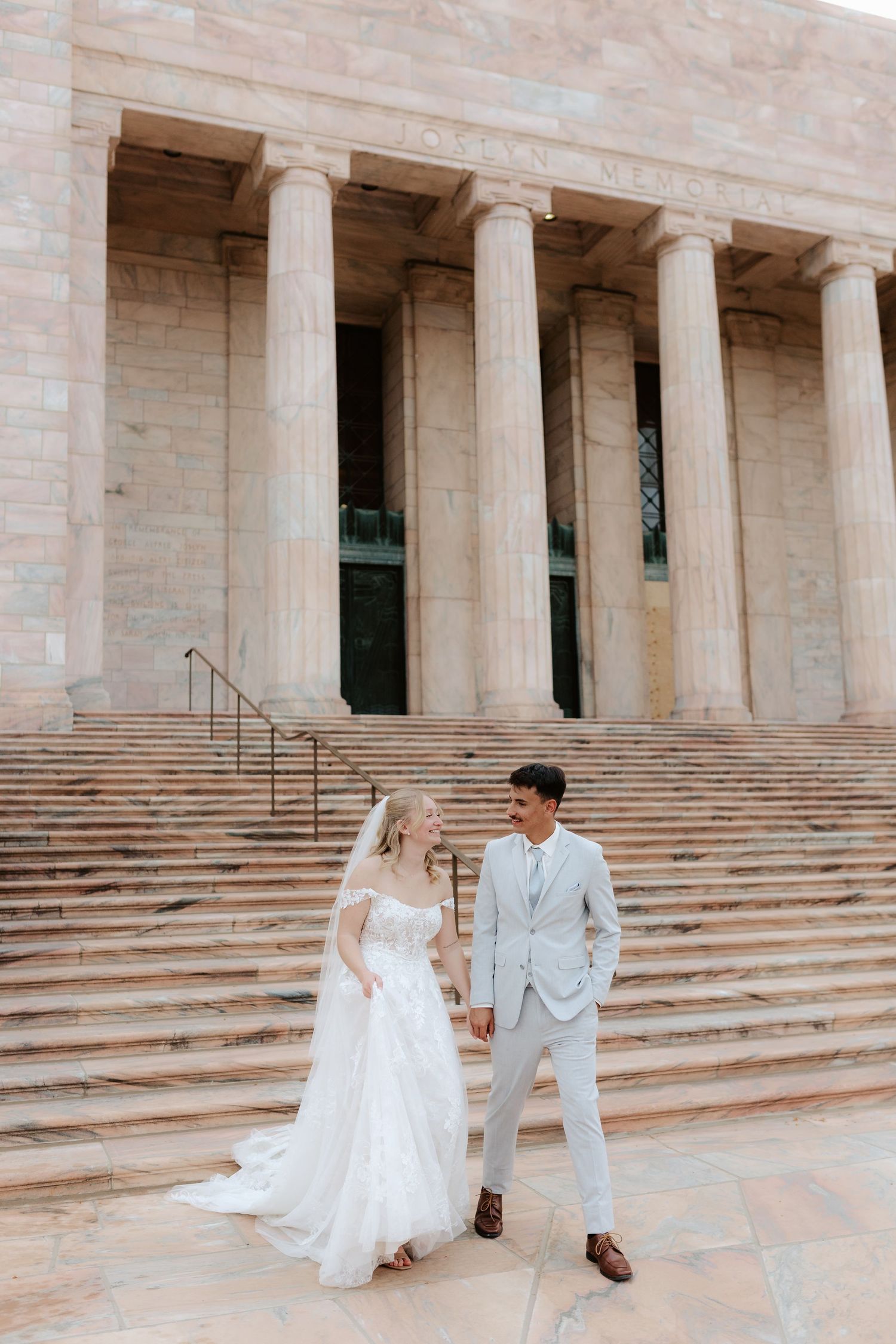A couple in wedding attire stands on grand stone steps in front of columned neoclassical architecture in Omaha Nebraska.