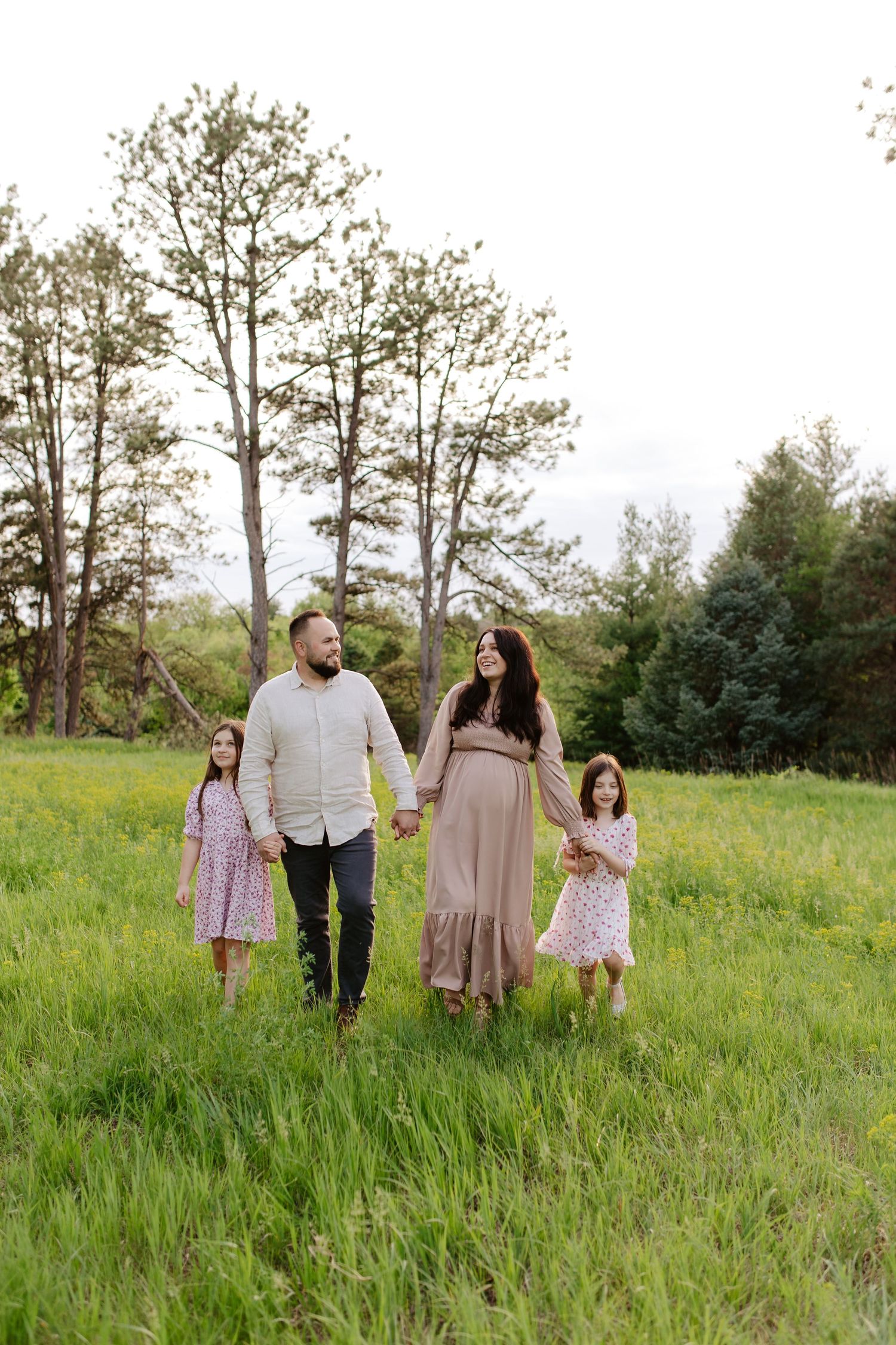 A family walks together holding hands in a green meadow with tall trees in the background on a summer day, maternity photoshoot.