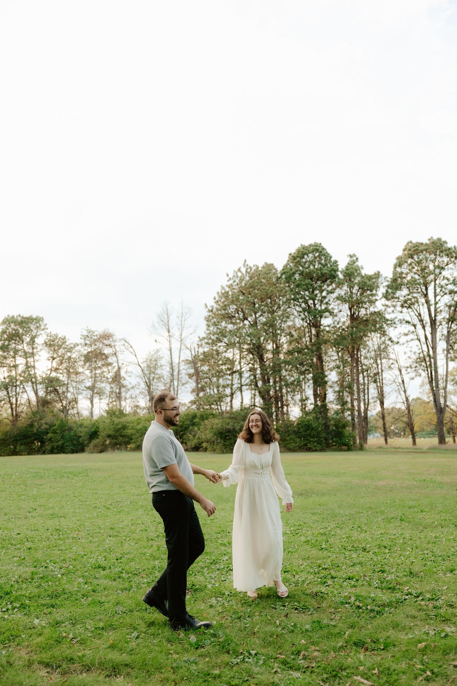 A couple dances together in a grassy field with autumn trees in the background during a romantic outdoor moment.