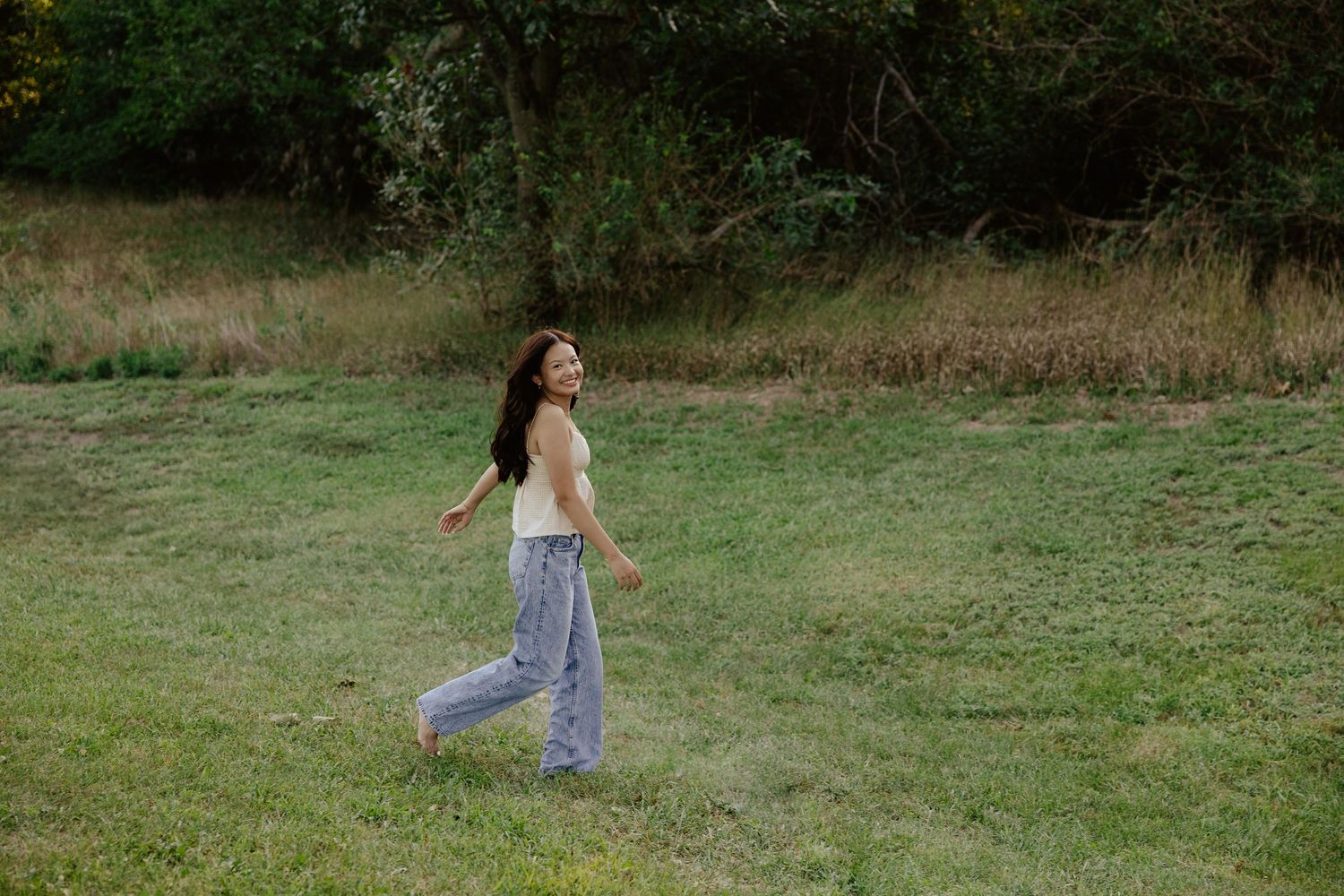 A girl walking through a grassy field in casual clothing at sunset for her senior photo session.