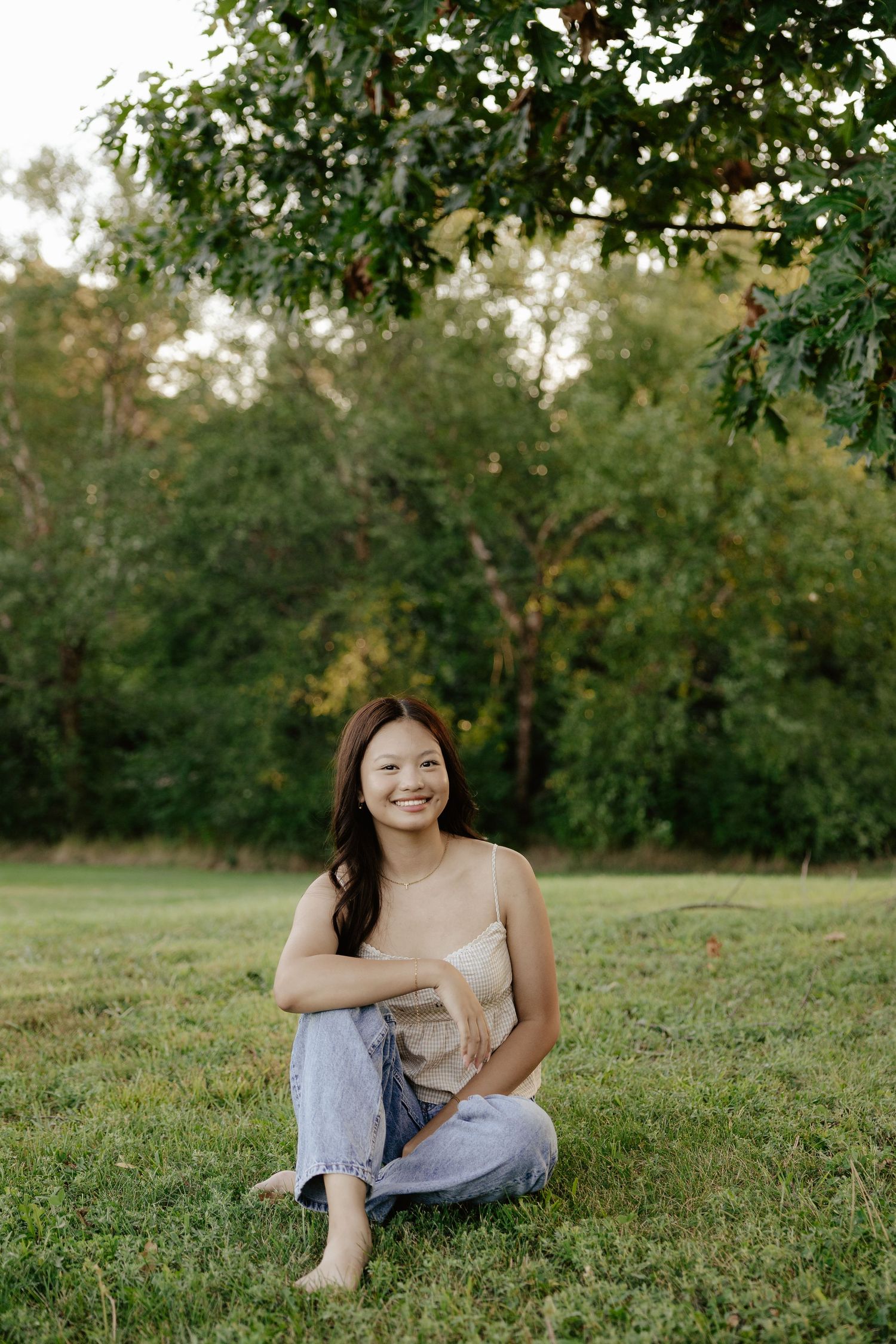 A girl in a beige top and light jeans sits on green grass with trees in the background during golden hour at Pioneers Park in Lincoln NE.