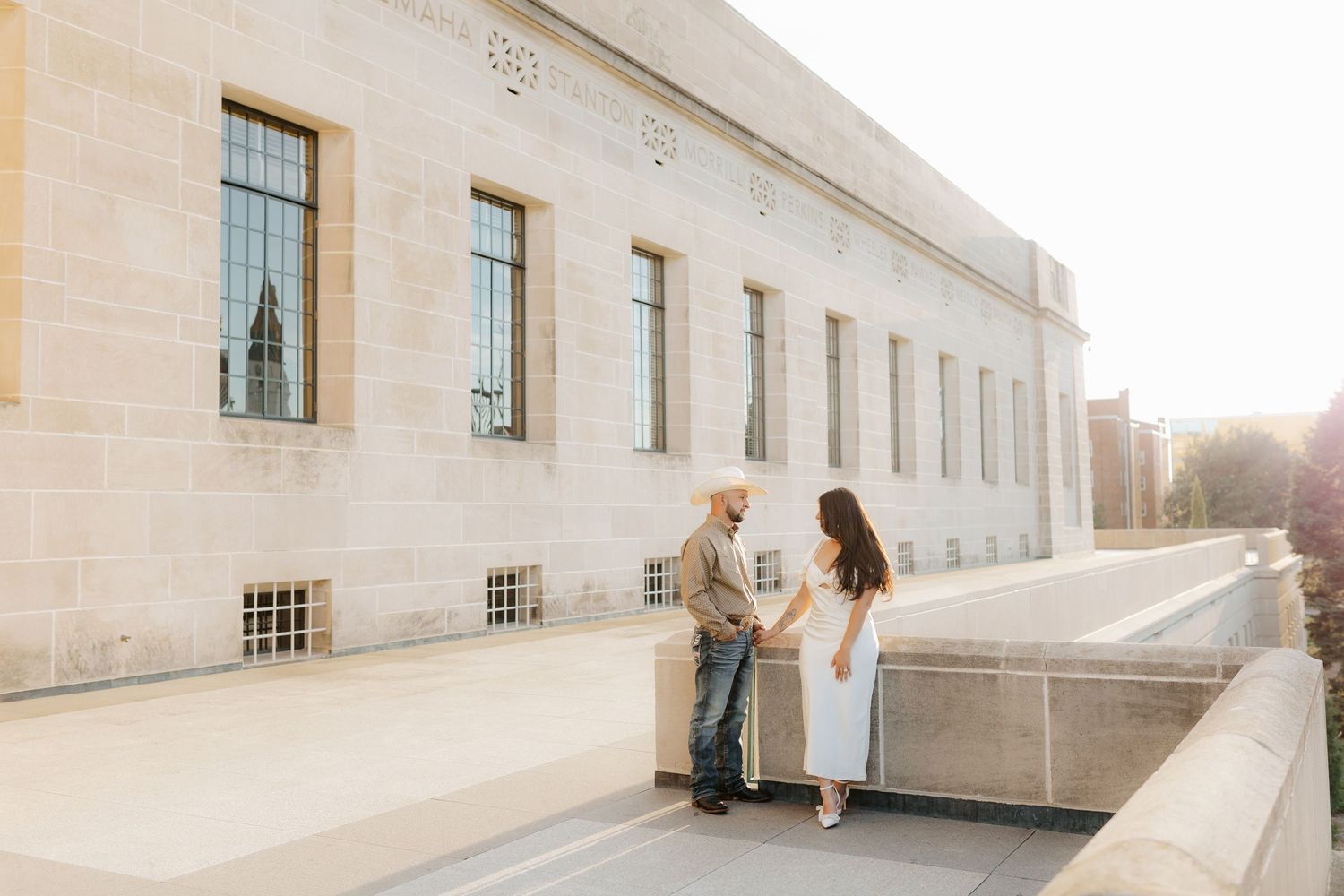 Sun flares behind the Nebraska State Capitol exterior as someone in a flowing white dress moves gracefully along the walkway.