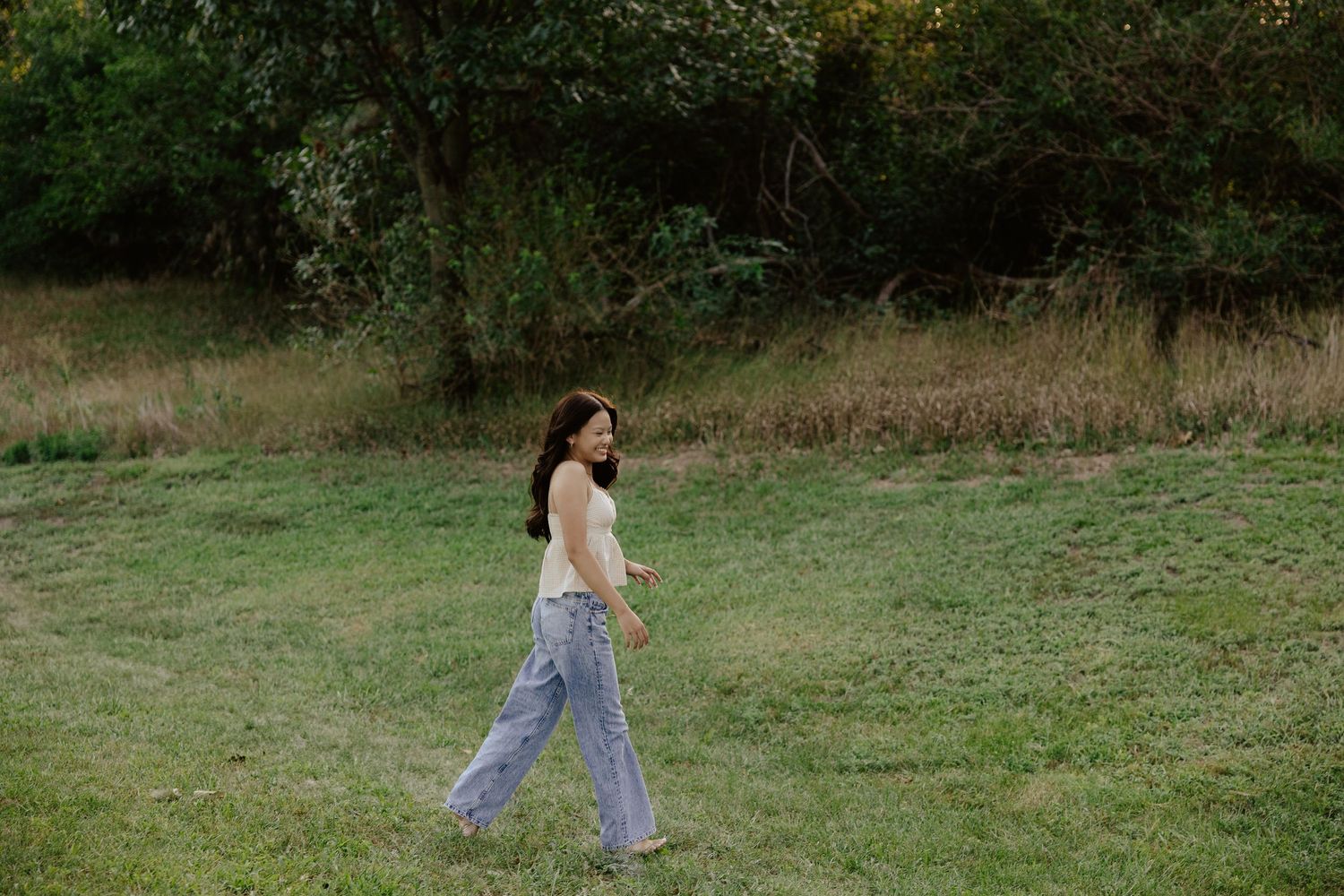 A girl walking through a grassy field in casual clothing at sunset for her senior photo session.
