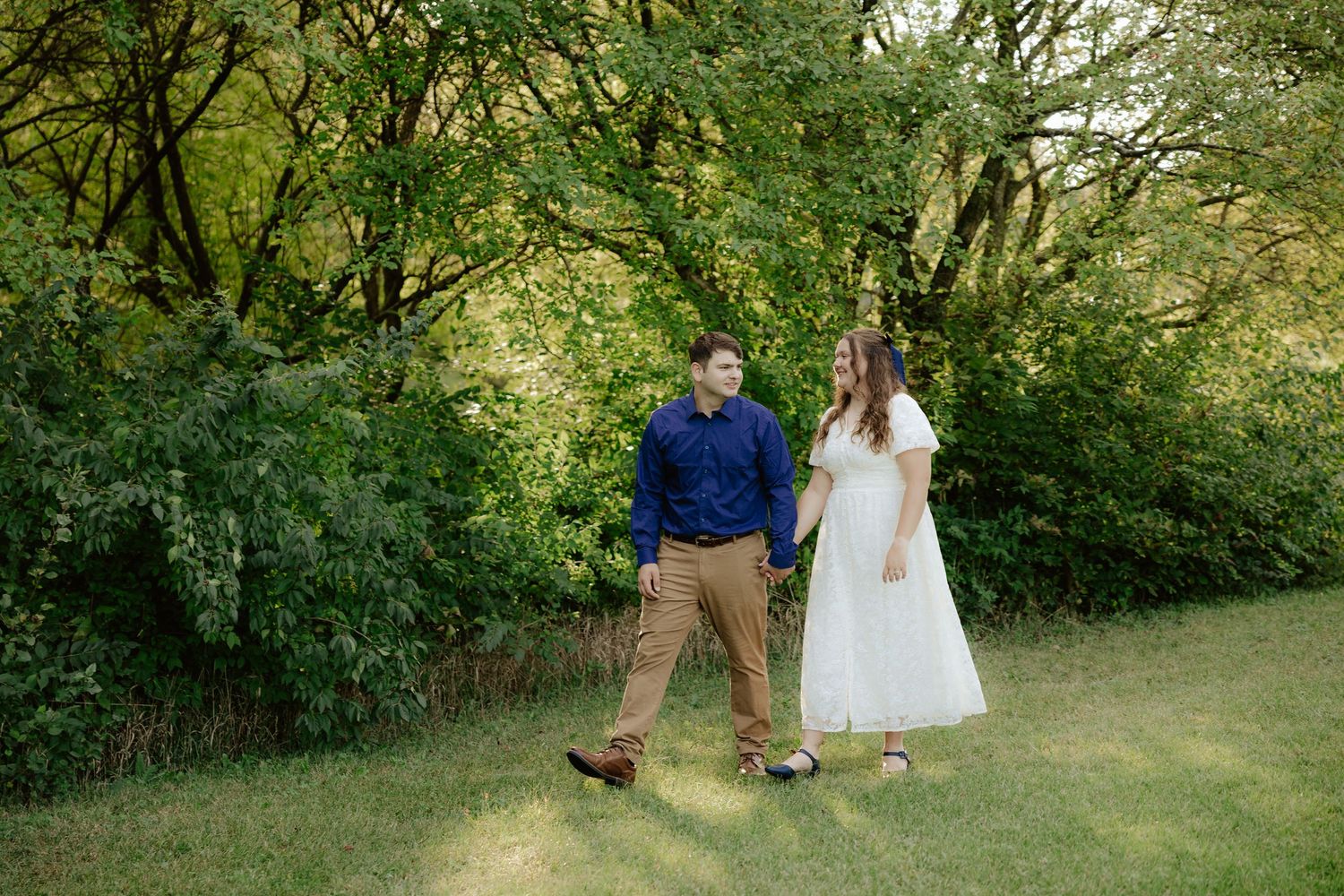 A couple walking hand in hand through lush green foliage on a summer afternoon.