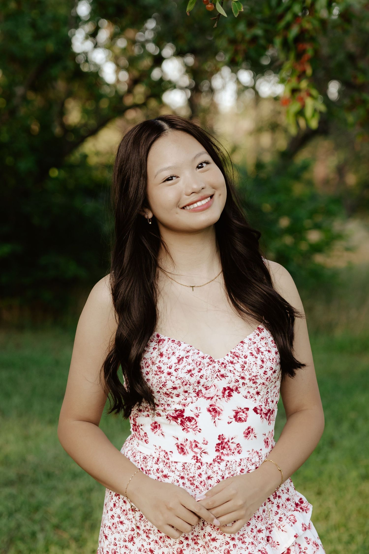 A girl in a white and red floral dress poses outdoors under blooming trees on a sunny day for her senior photos.