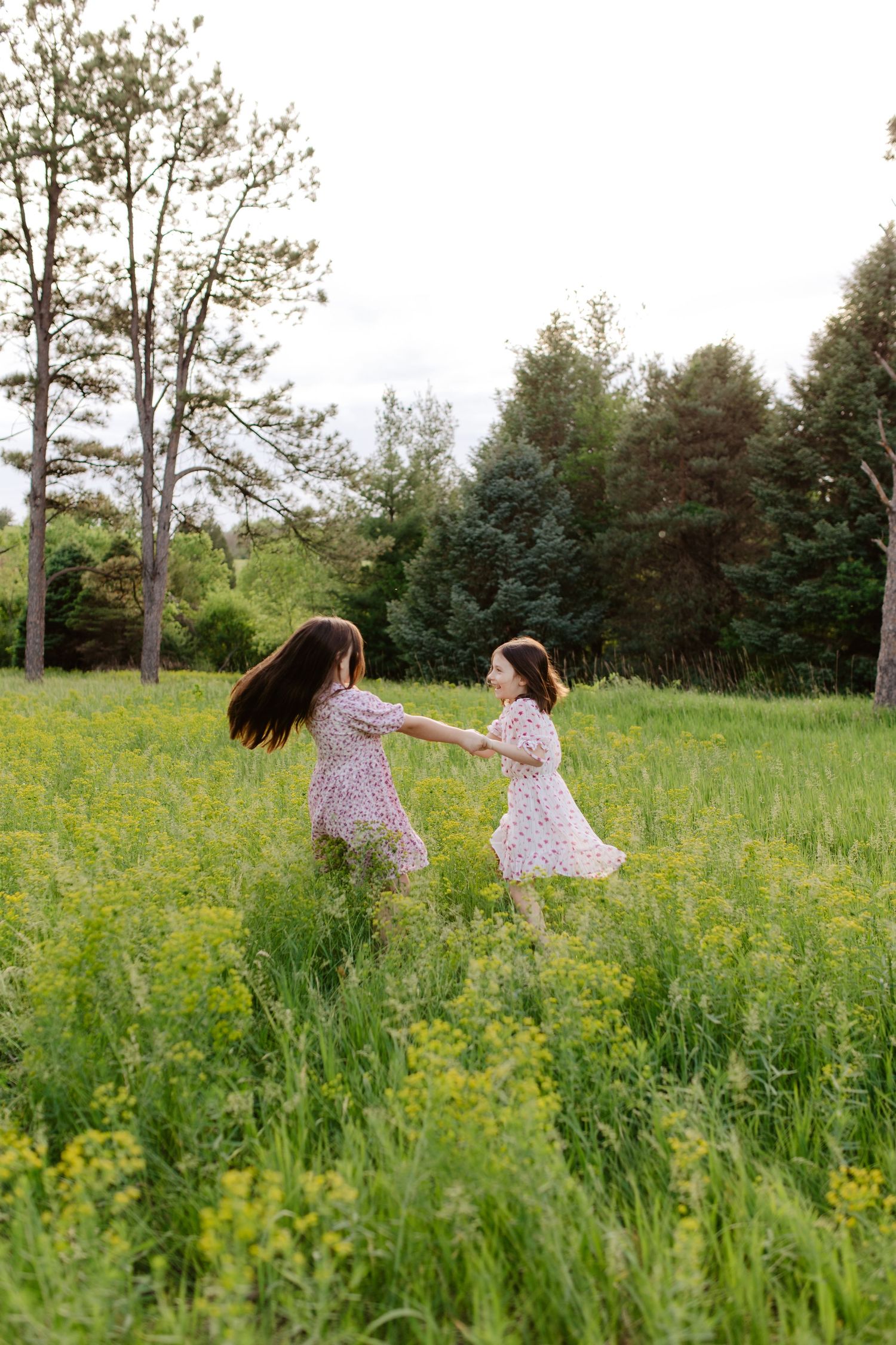 Two sisters in flowy dresses run together through a sunlit meadow filled with yellow wildflowers.