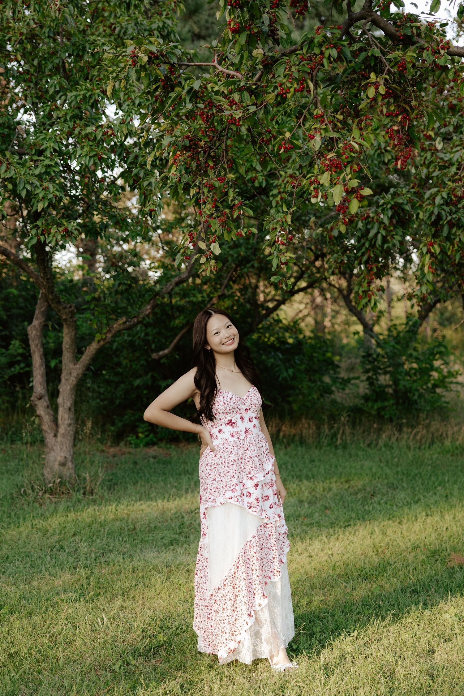 A girl in a white and red floral dress poses outdoors under blooming trees on a sunny day for her senior photos.
