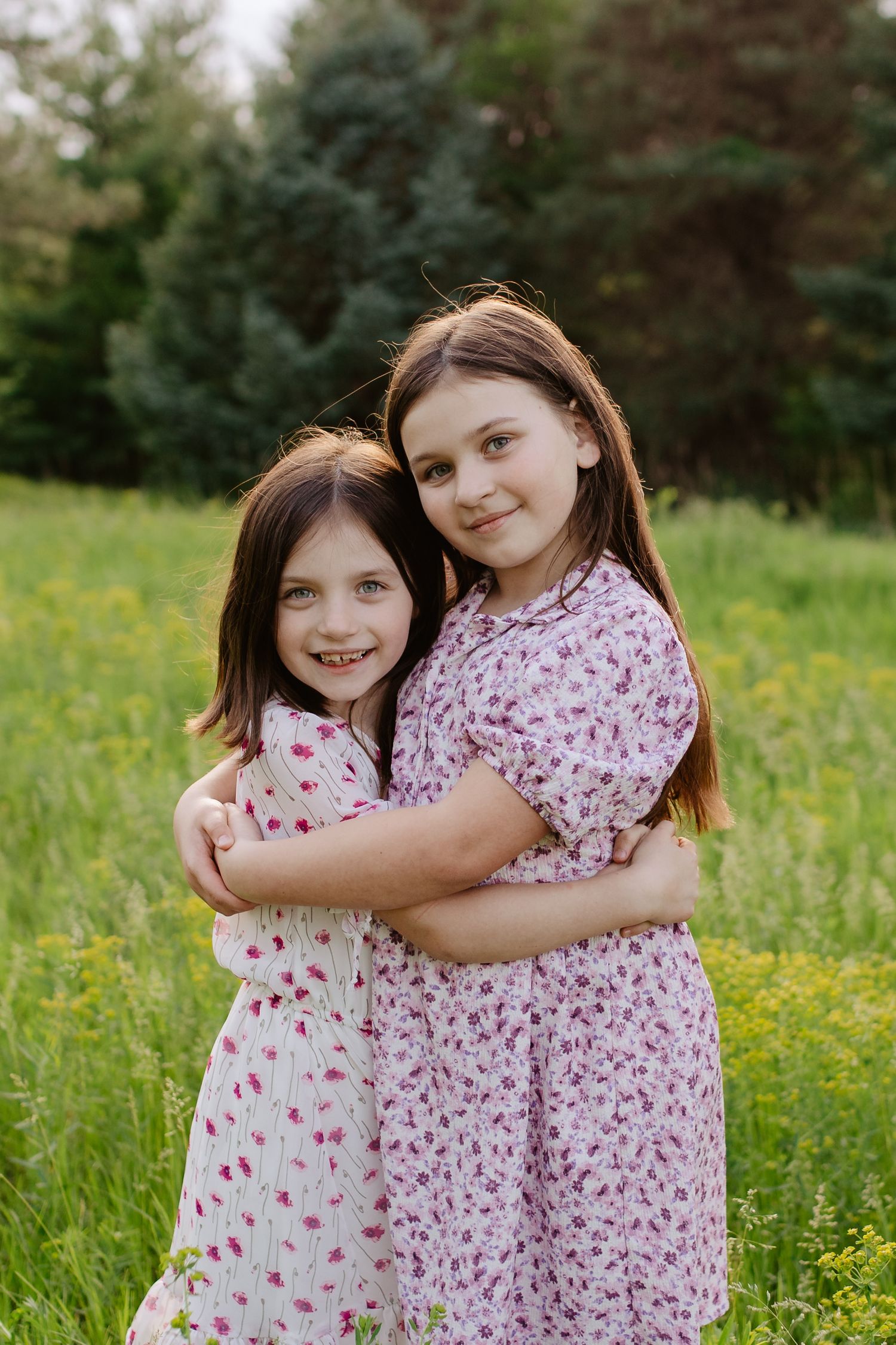 Two children in floral dresses sit together in a grassy meadow with wildflowers on a sunny day.