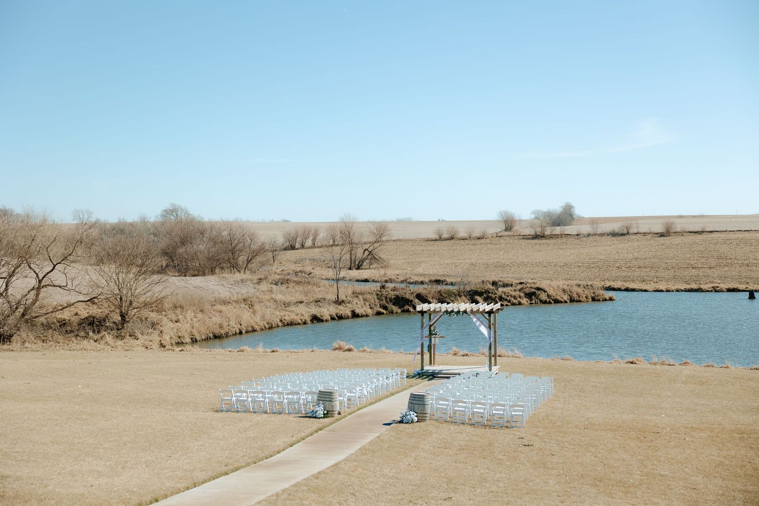 Outdoor wedding altar with wooden cross, white flowing fabric, and blue floral decor beside a serene pond at Sparks Barn in Nebraska
