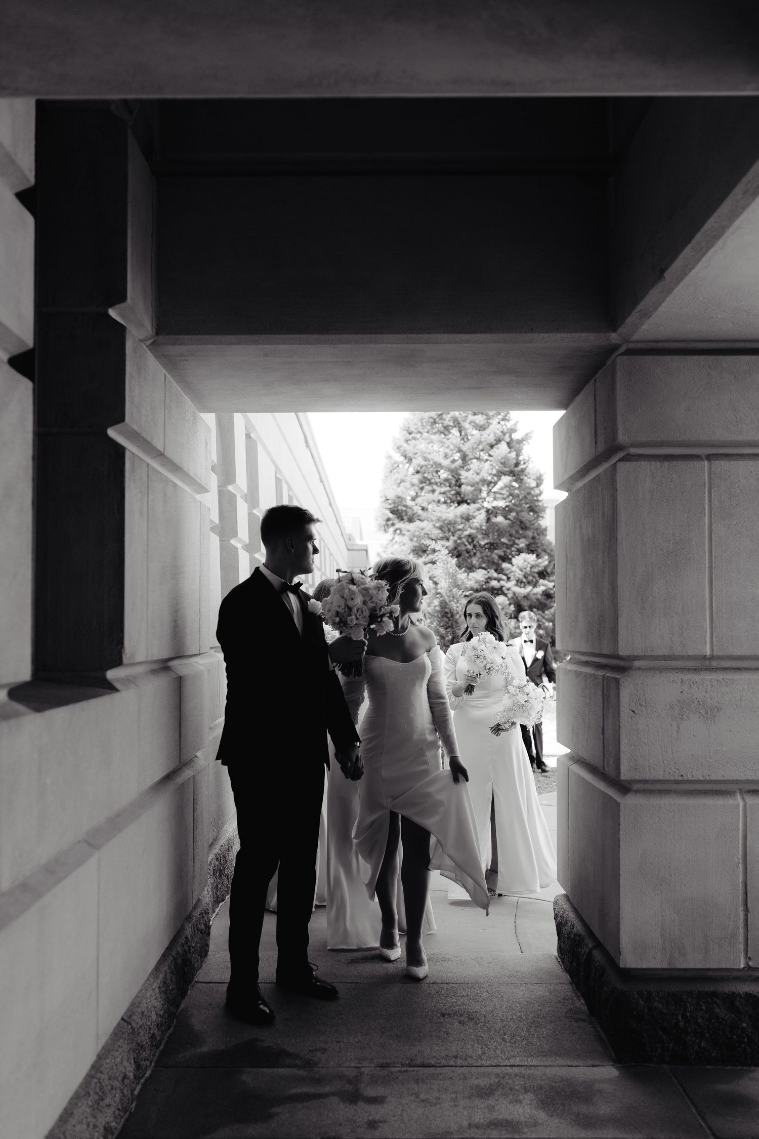 Black and white photo of a bride and groom walking under an archway, with wedding party members nearby at the Nebraska State Capitol