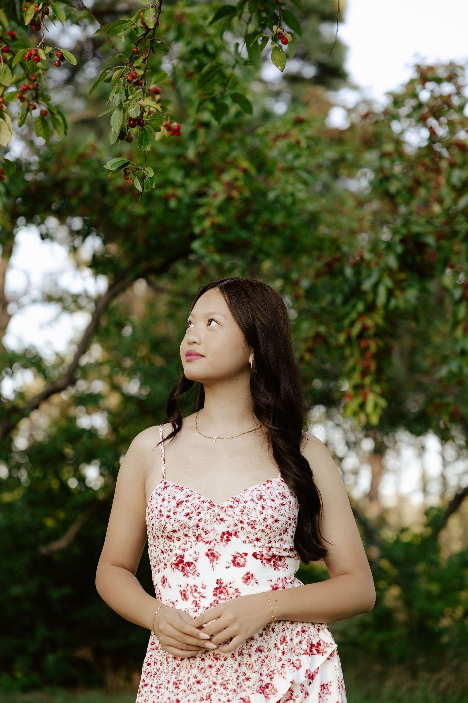 A girl in a white and red floral dress poses outdoors under blooming trees on a sunny day for her senior photos.