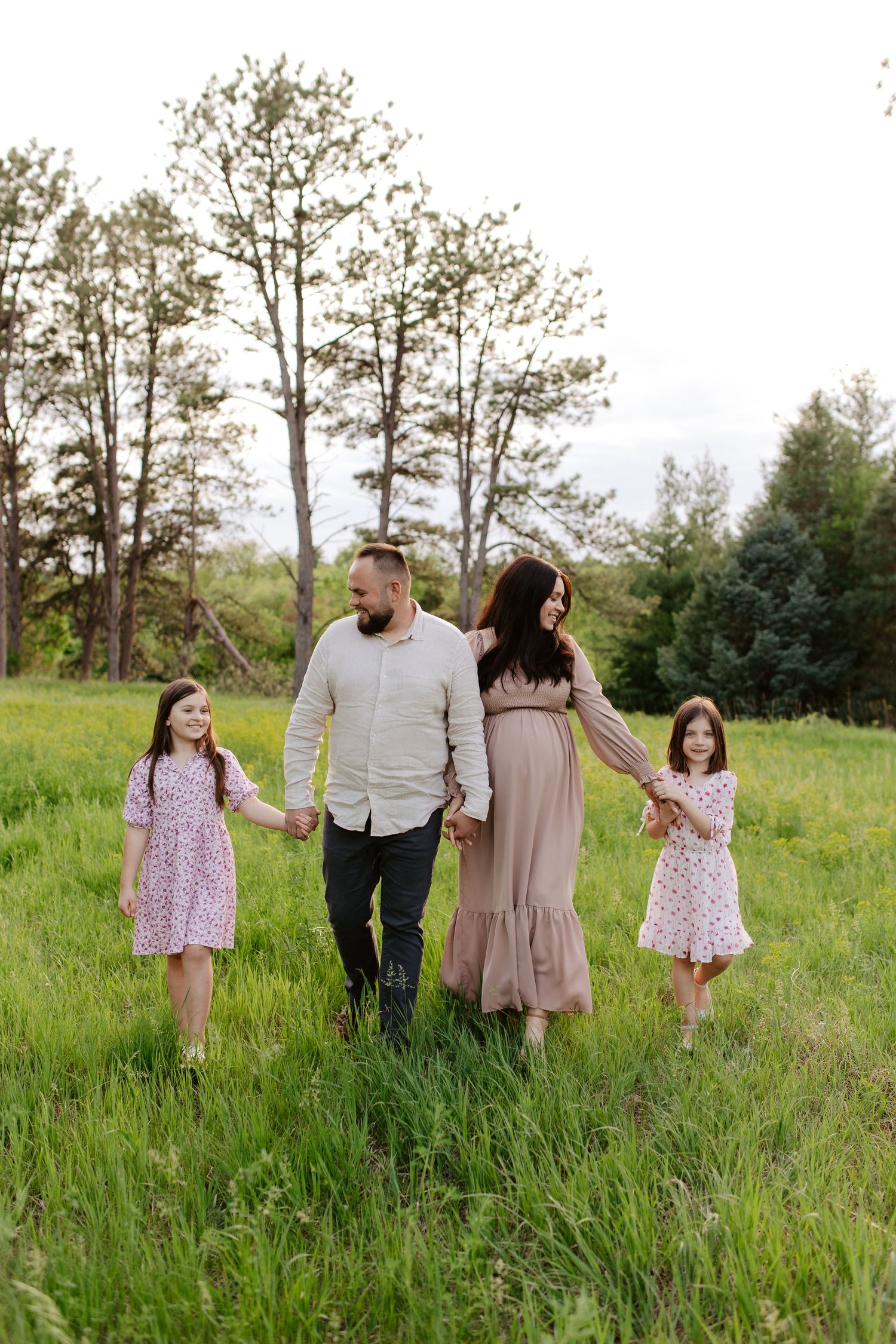 A family walks together holding hands in a green meadow with tall trees in the background on a summer day, maternity photoshoot.