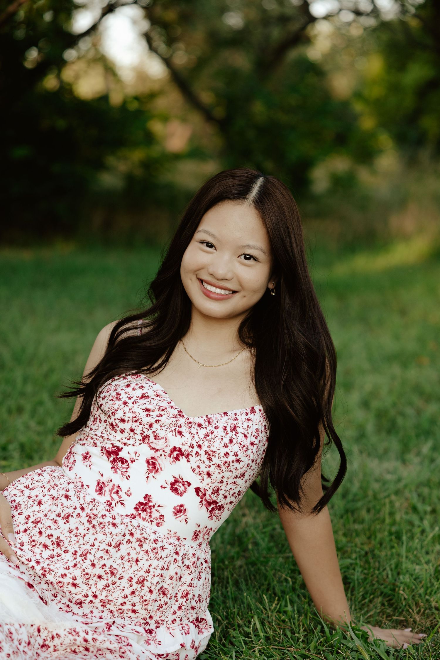 A girl in a red and white floral dress sits on green grass during sunset for her senior photo session.