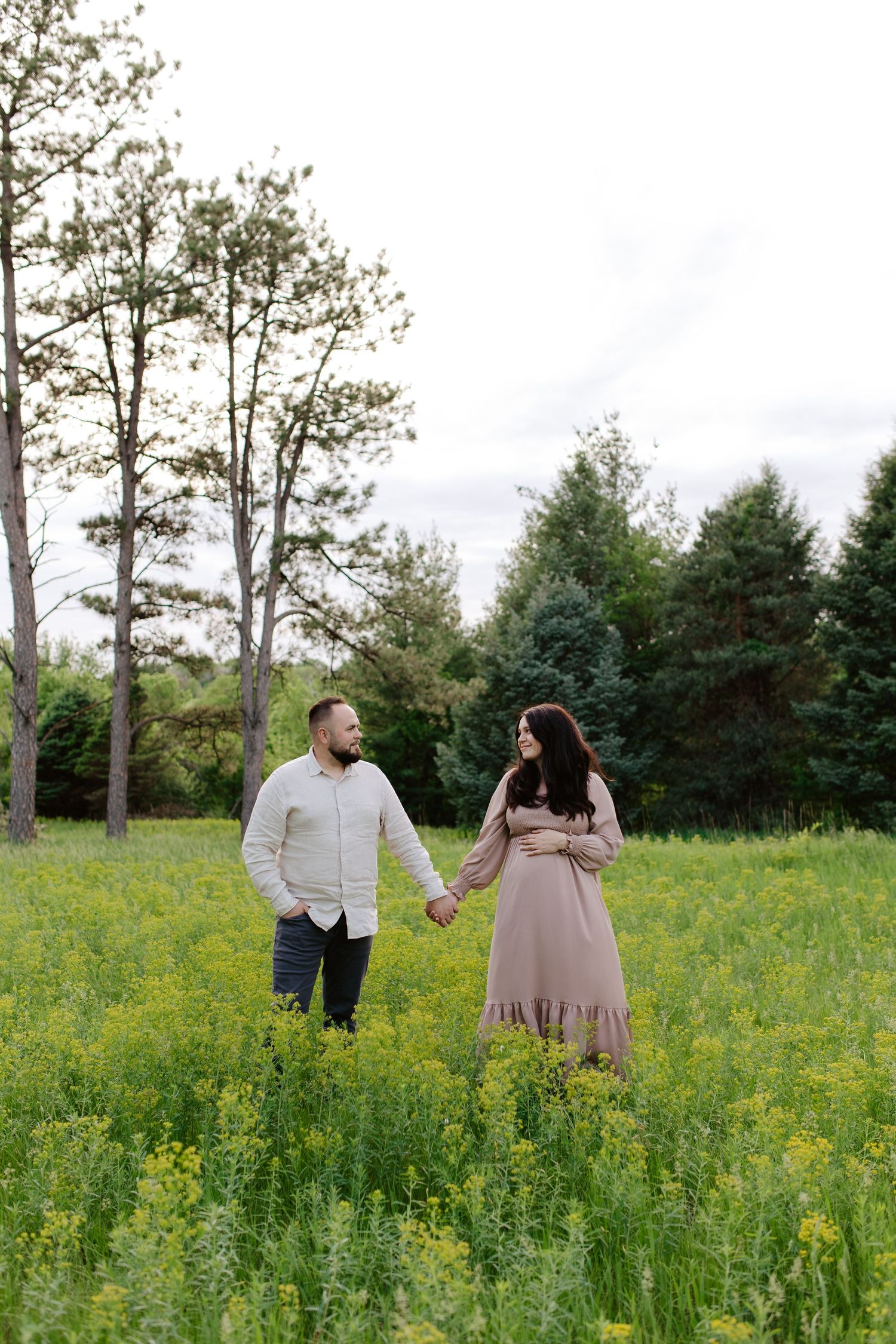 Two people hold hands while standing in a lush meadow of yellow wildflowers with evergreen trees in the background.