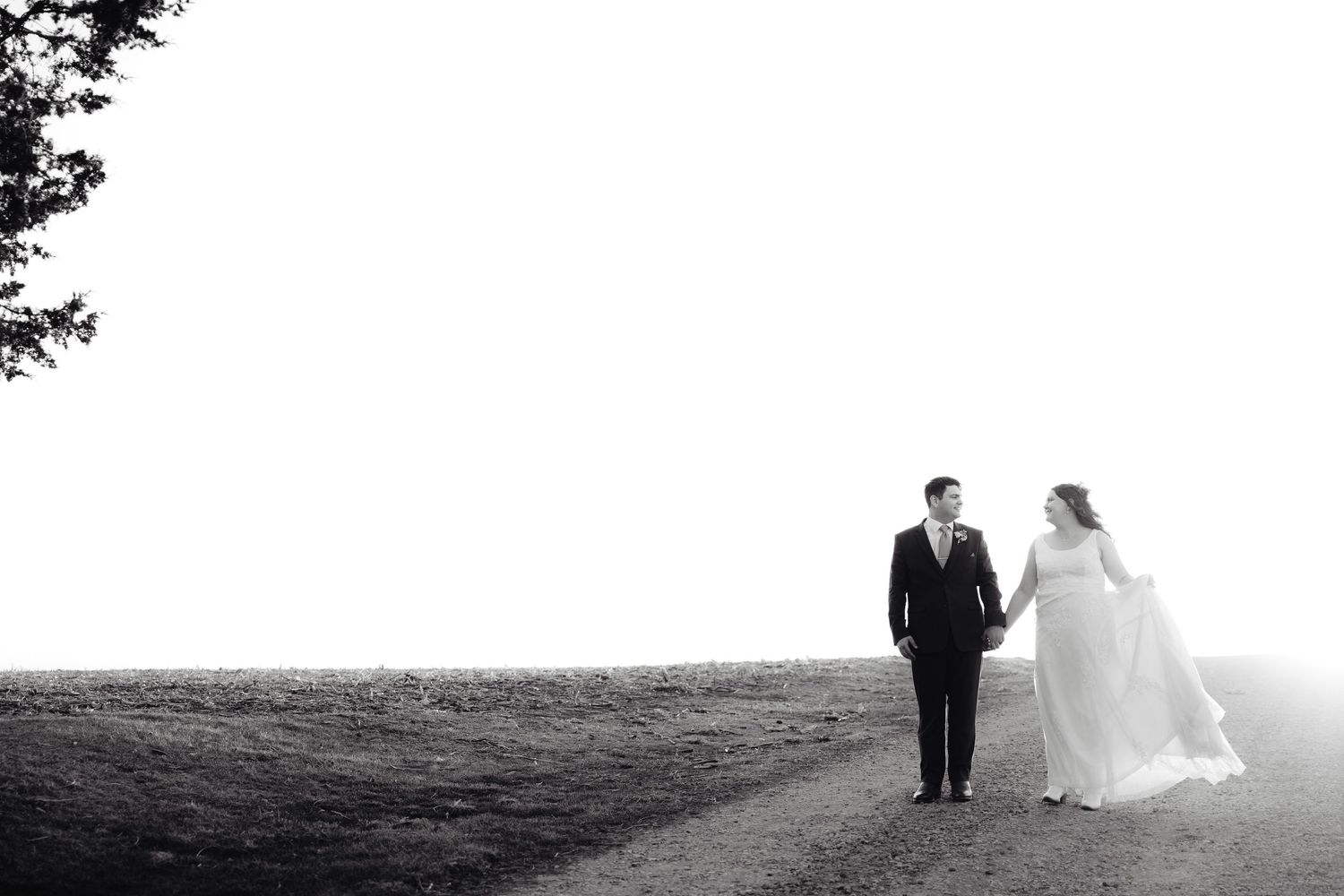 Black and white photo of a bride and groom holding hands while walking on a path through an open field.