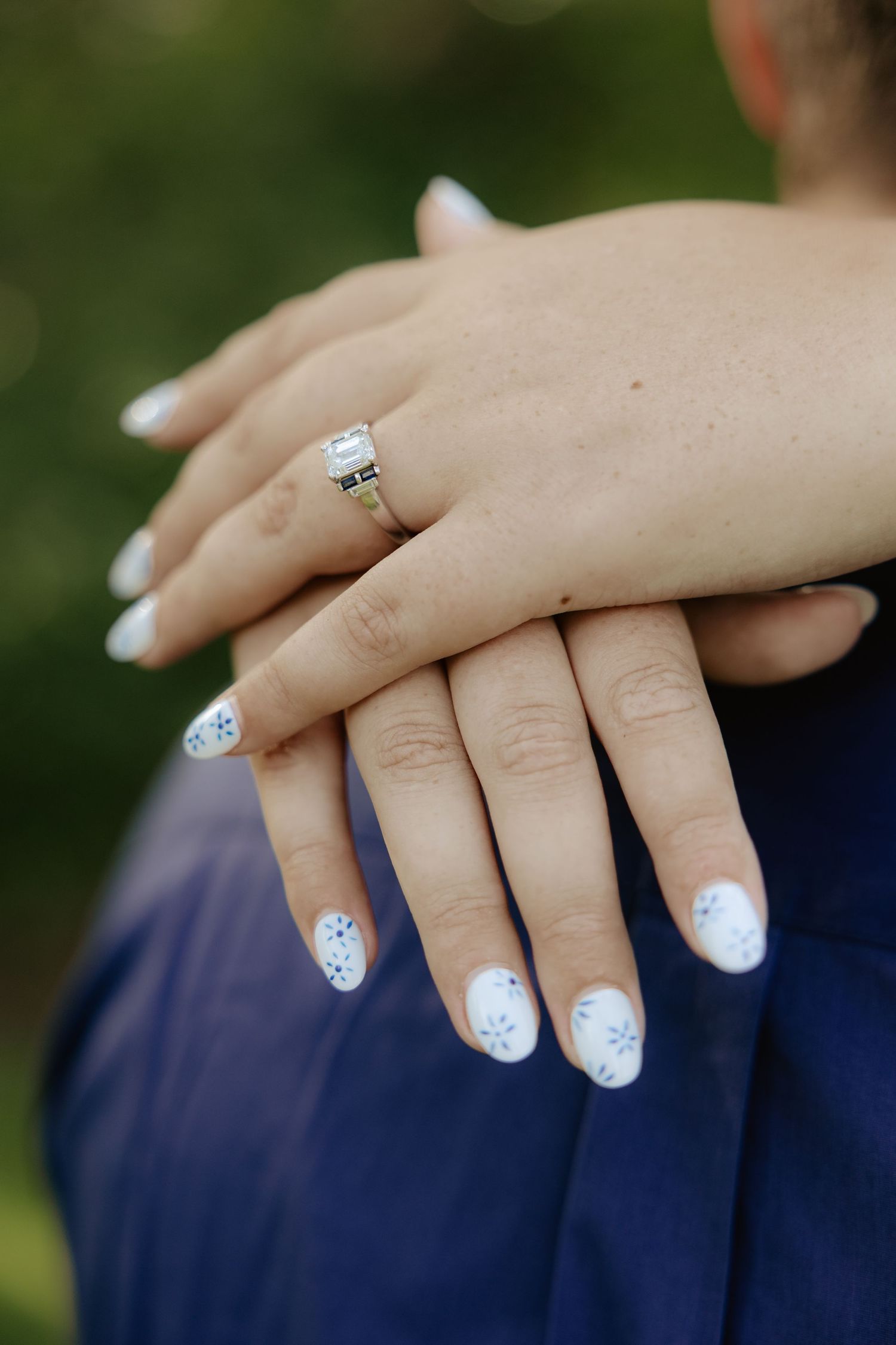 Close-up series of manicured hands with white nail polish and delicate blue floral designs wearing an engagement ring.