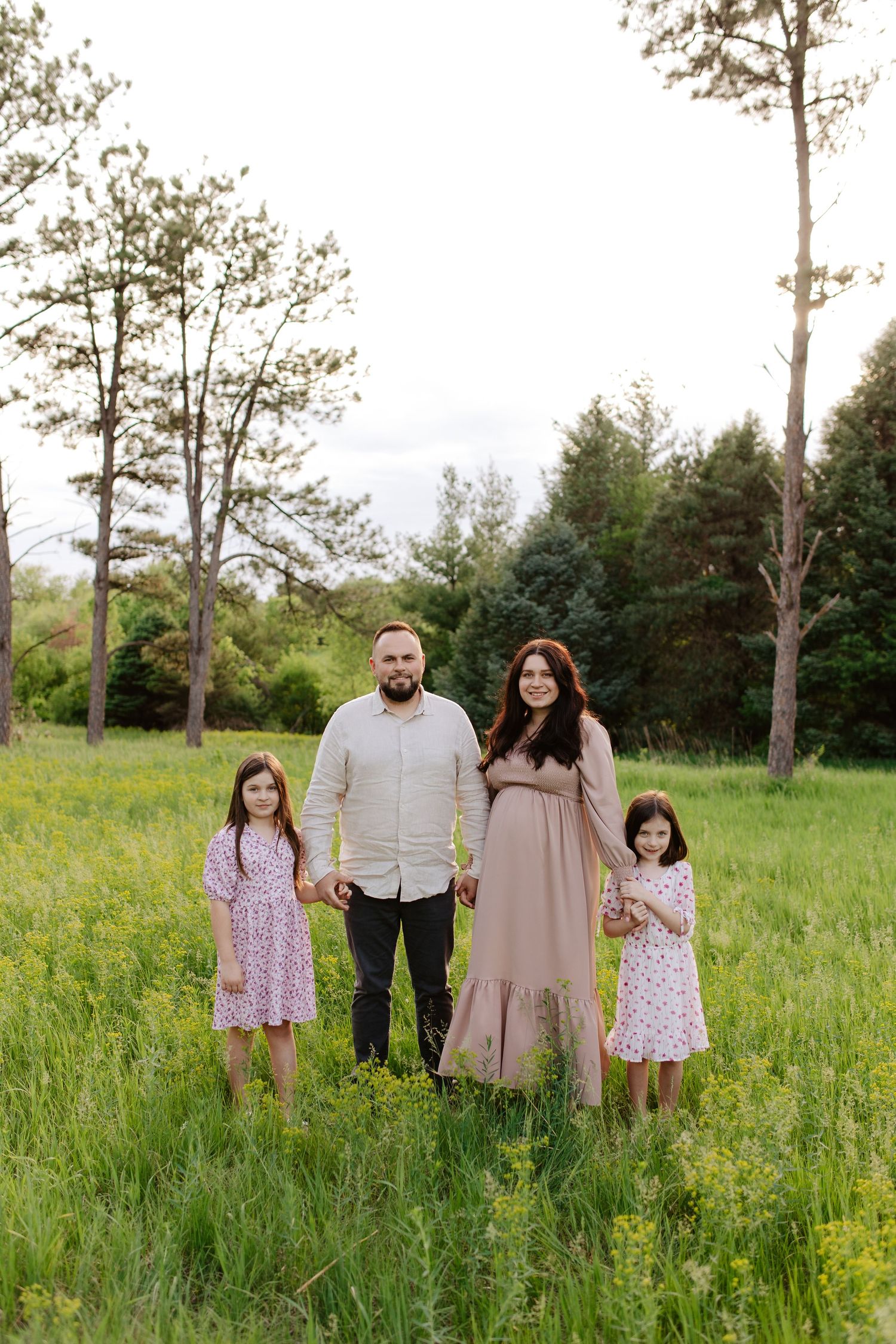 A family poses together holding hands in a green meadow with tall trees in the background on a summer day, maternity photoshoot.