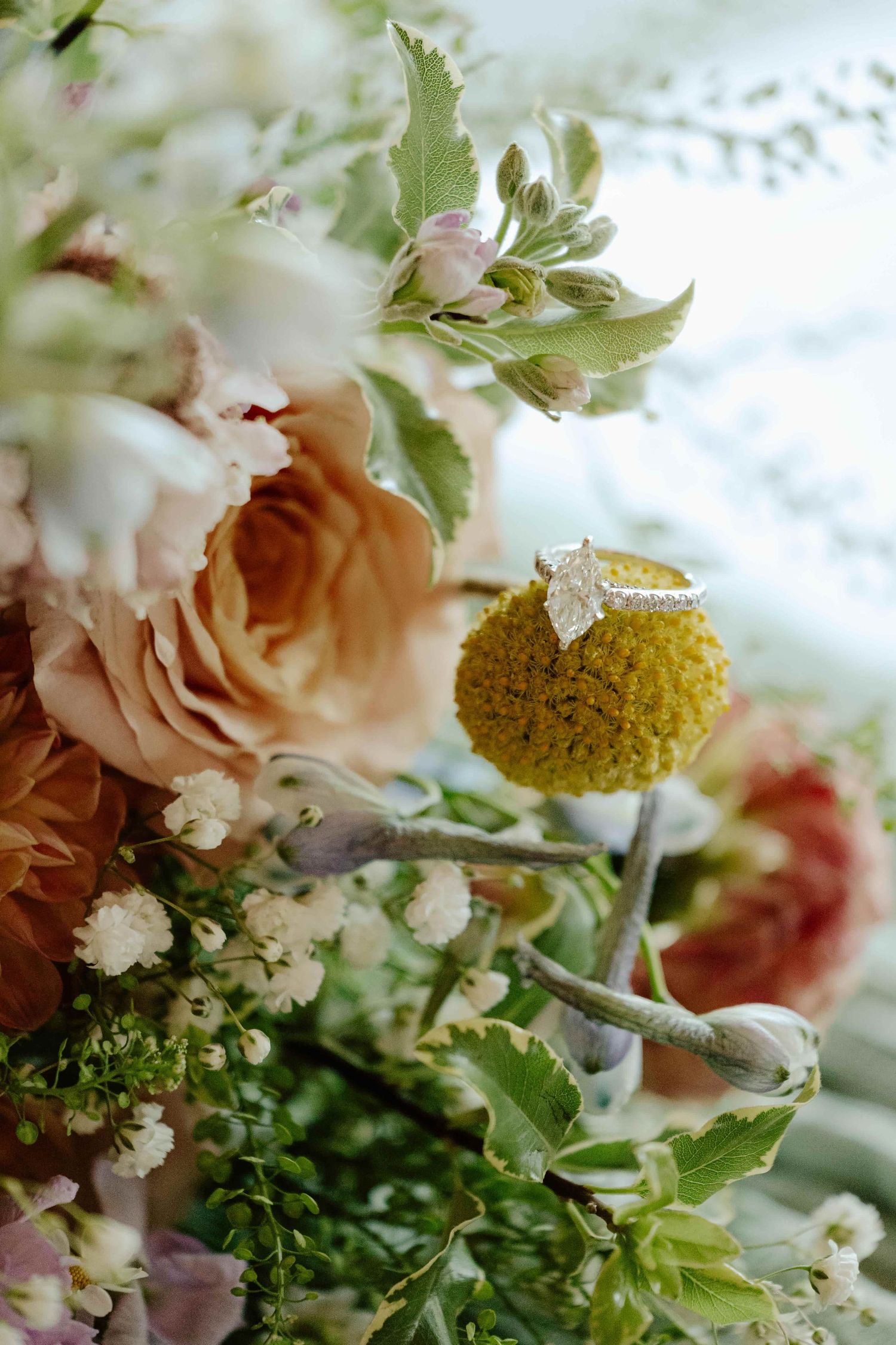 A diamond engagement ring resting on a yellow craspedia bloom surrounded by peach roses and lush greenery in a bridal bouquet.