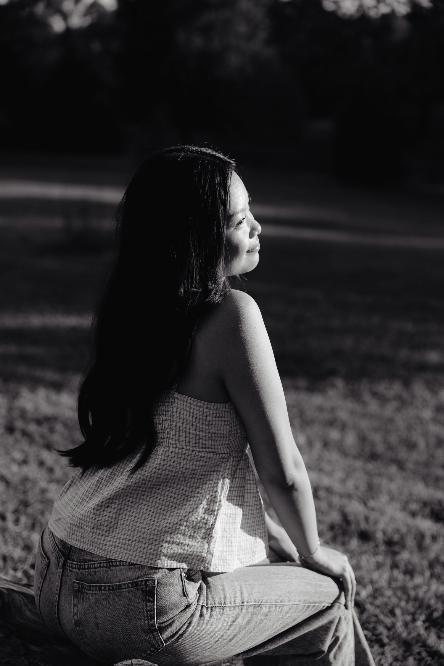 A girl in a beige top and light jeans sits on a log with trees in the background during golden hour at Pioneers Park in Lincoln NE.