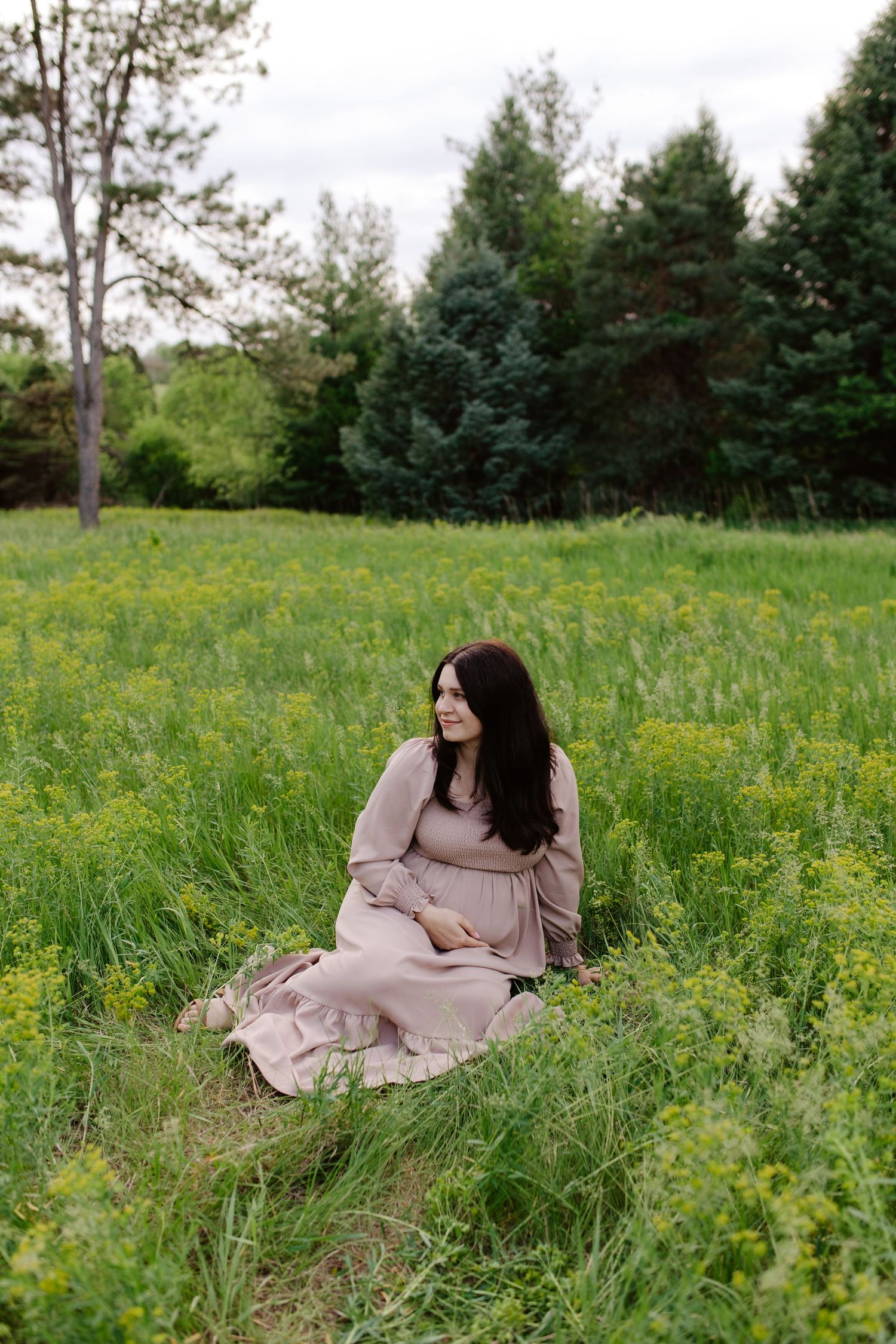 A woman in a flowing blush dress sits peacefully in a wildflower meadow with evergreen trees in the background.