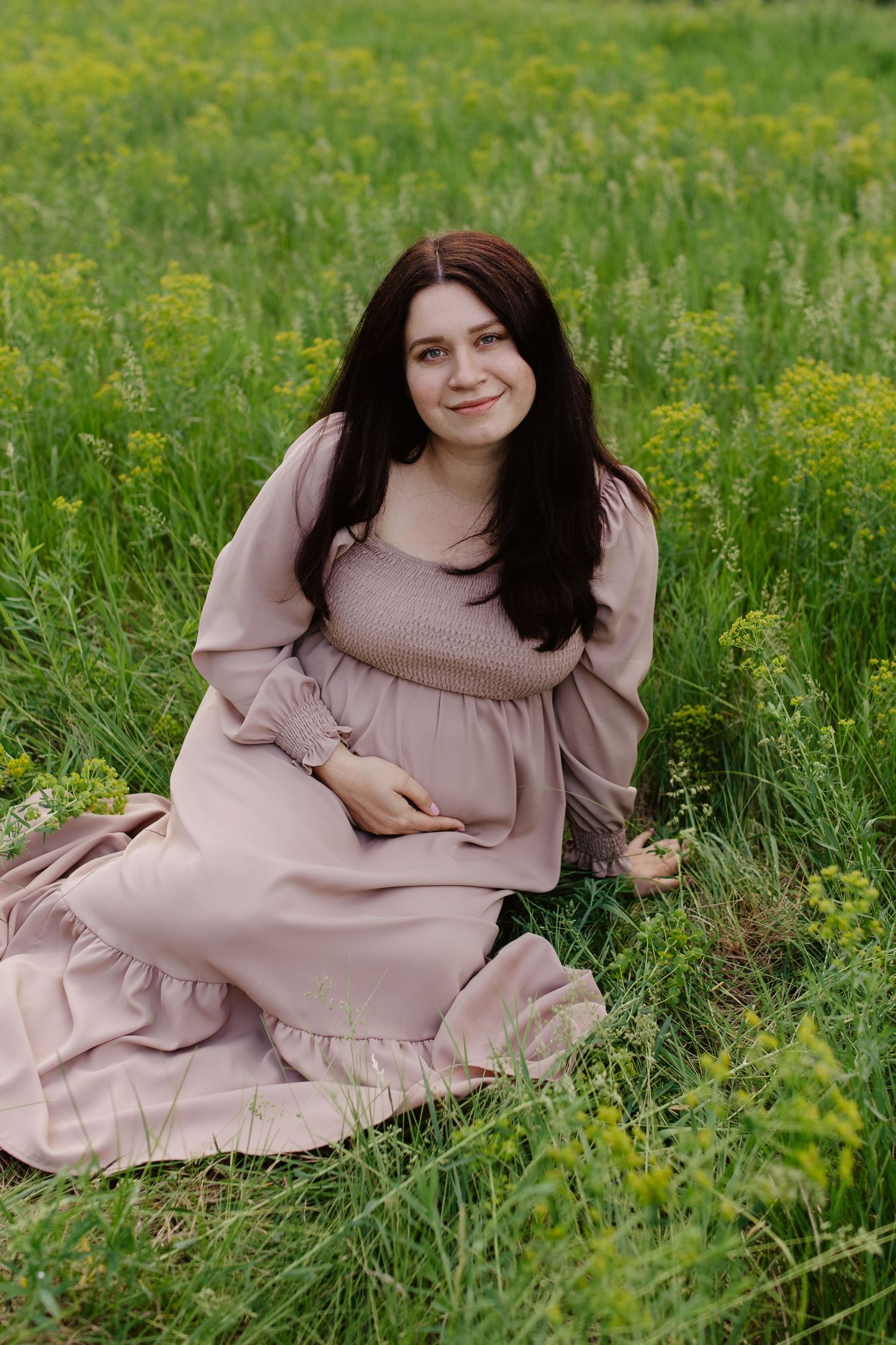 Woman in flowing pink dress sits gracefully in a meadow filled with yellow wildflowers on a sunny day.