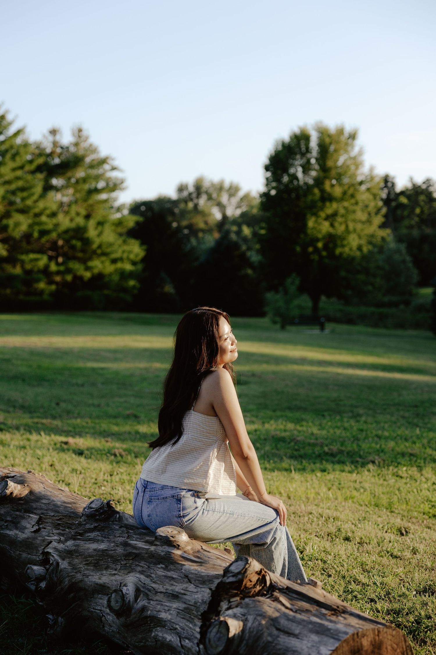 A girl in a beige top and light jeans sits on a log with trees in the background during golden hour at Pioneers Park in Lincoln NE.
