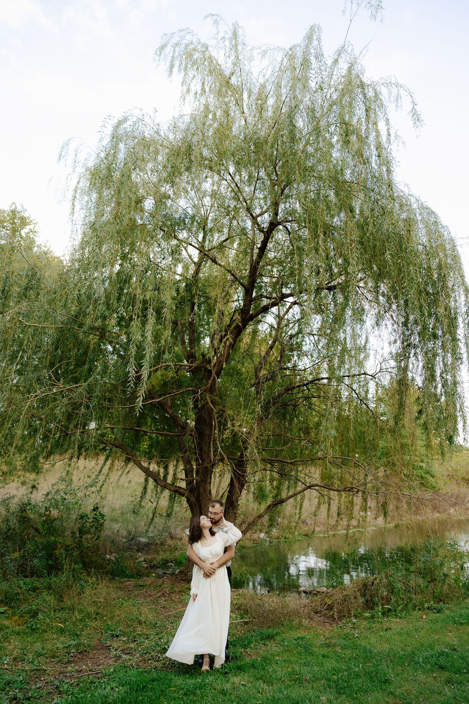 A couple embraces under a weeping willow tree by a tranquil pond in a romantic outdoor setting.