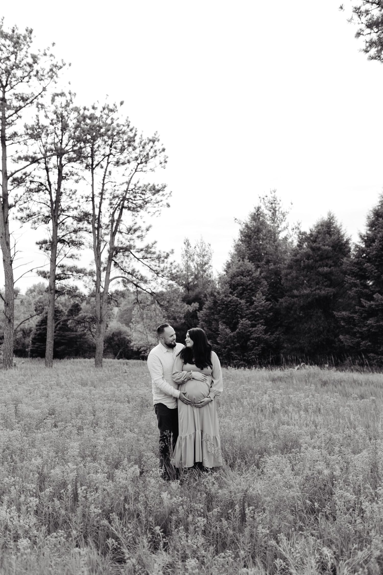 A couple shares a tender moment together in a wildflower meadow surrounded by tall trees in black and white photography.
