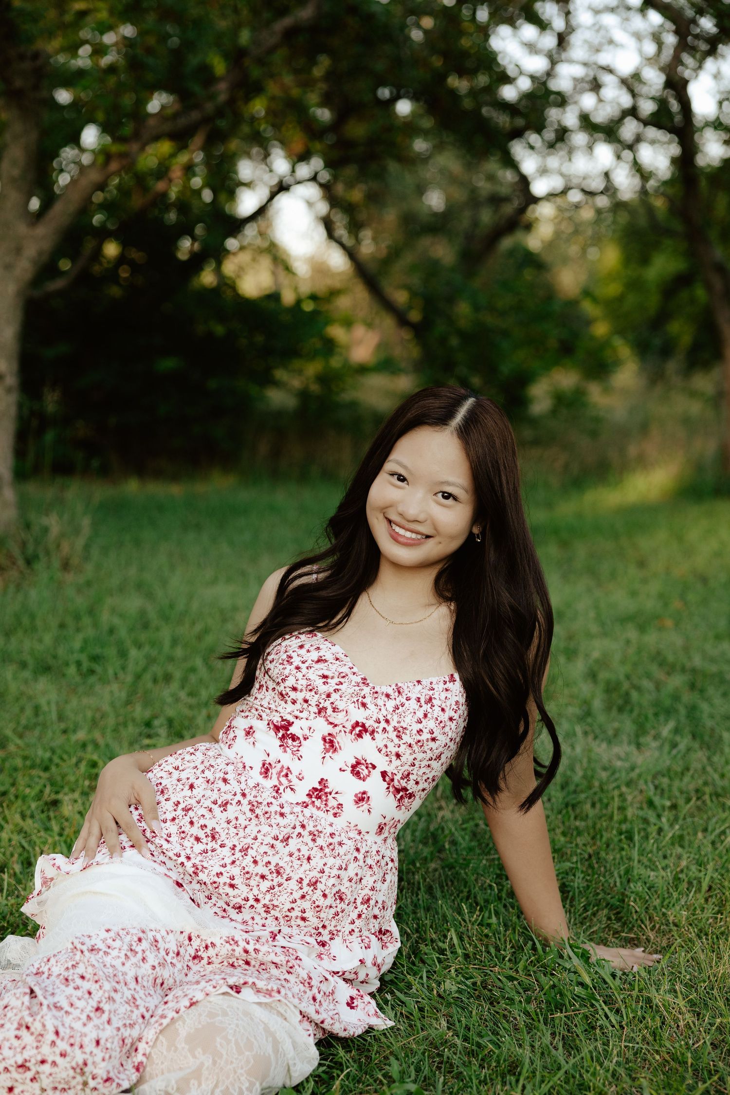 A girl in a red and white floral dress sits on green grass during sunset for her senior photo session.