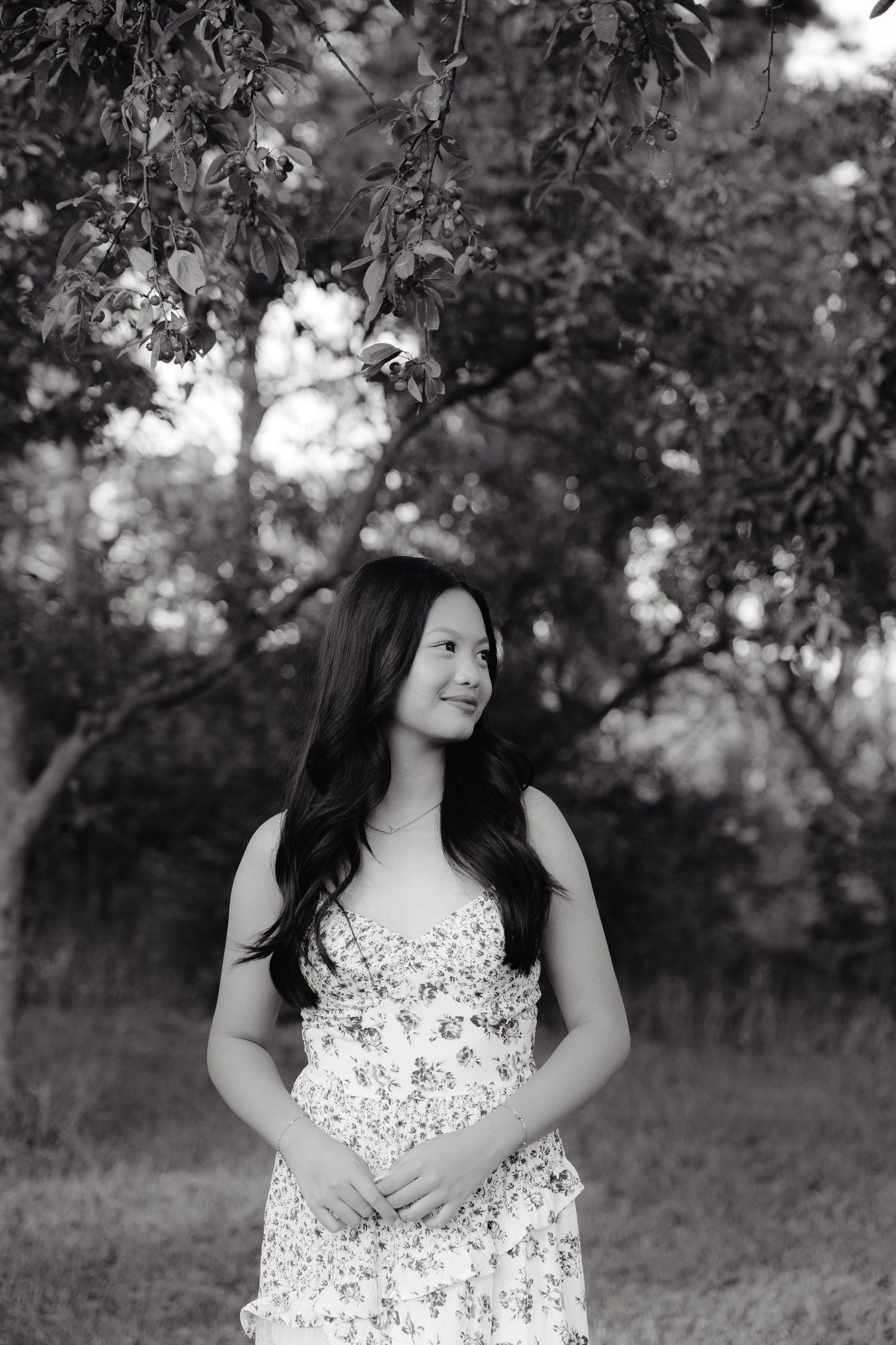 A girl in a white and red floral dress poses outdoors under blooming trees on a sunny day for her senior photos.