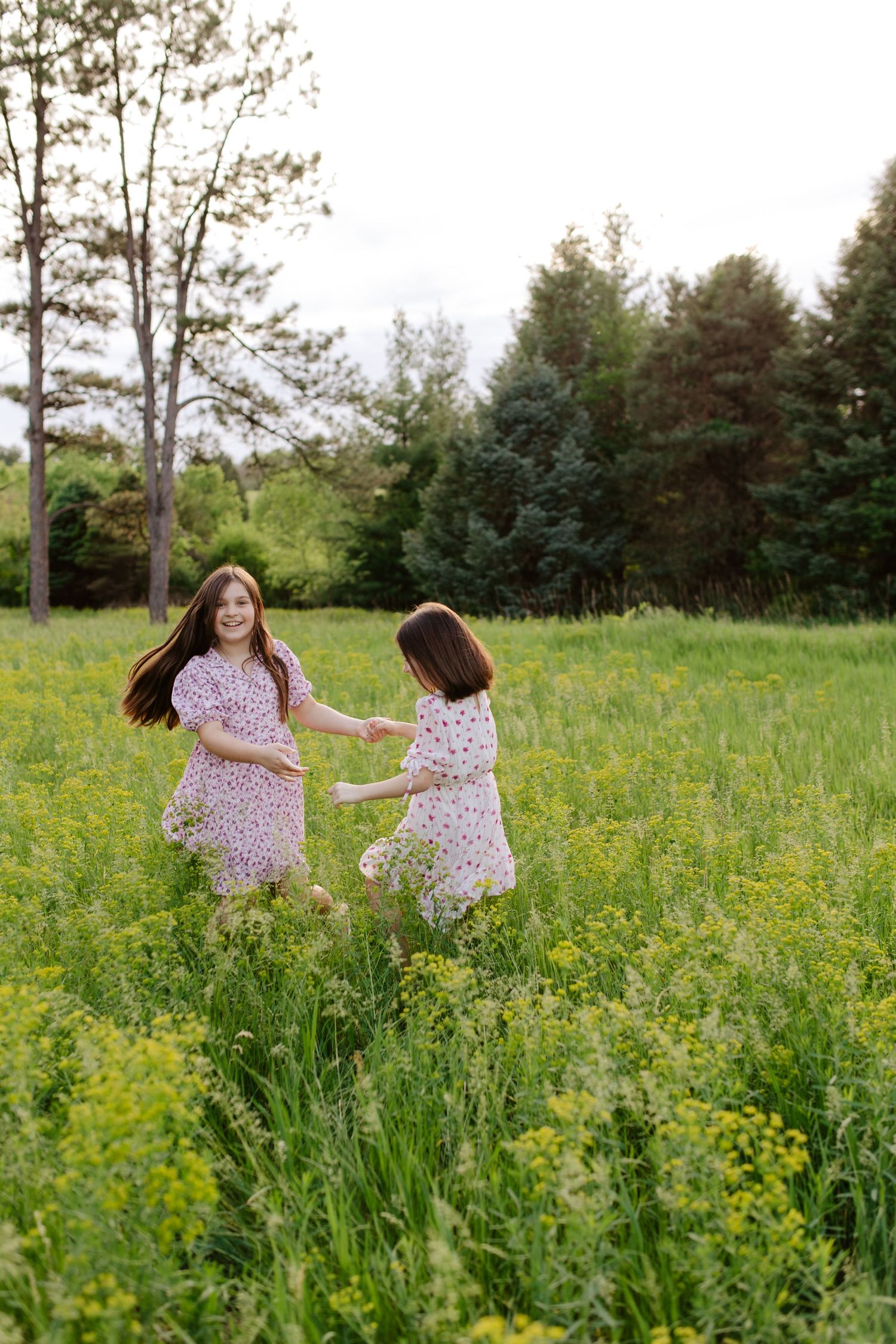Two sisters in flowy dresses run together through a sunlit meadow filled with yellow wildflowers.