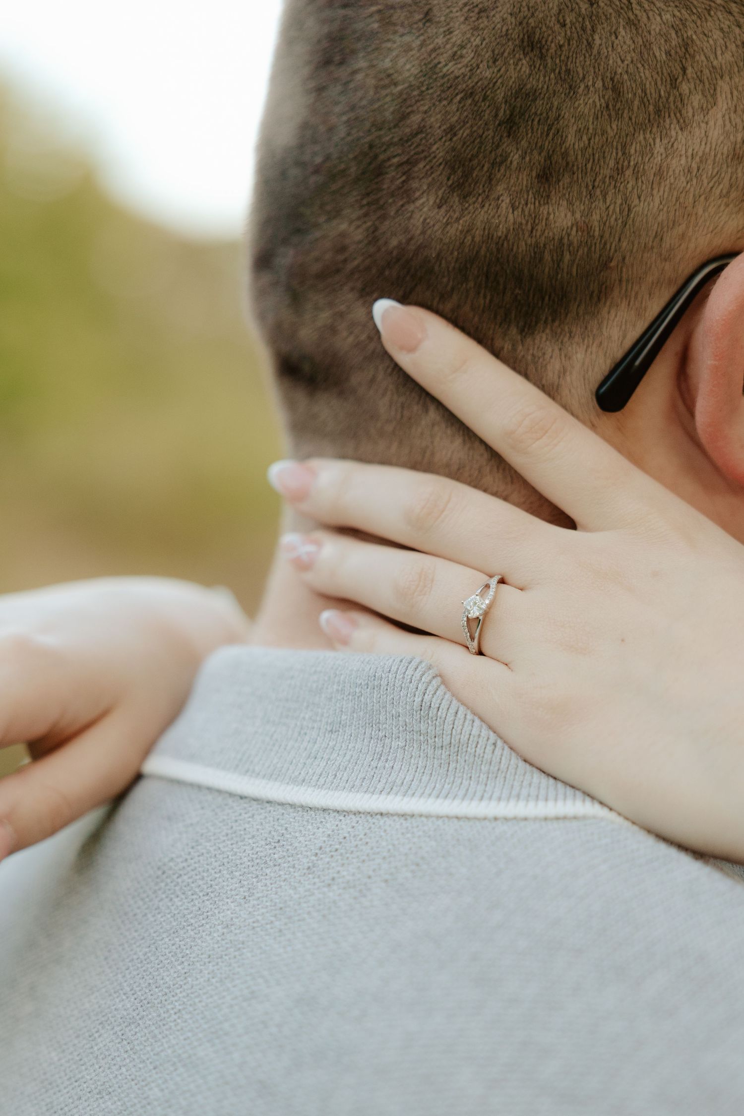 Close-up view of a hand wearing an engagement ring resting on a gray sweater shoulder.