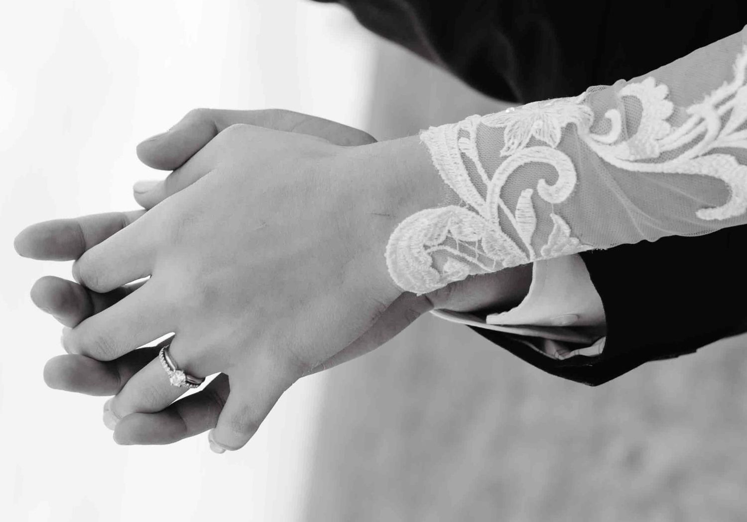 Black and white photo of a couple holding hands, showing wedding rings and lace bridal sleeve detail.