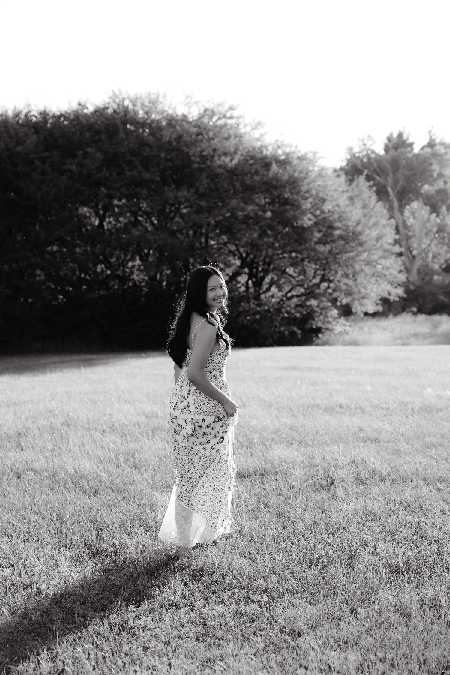 A girl in a white floral dress twirls playfully in a sunlit meadow surrounded by green trees at Pioneers Park in Lincoln Nebraska.