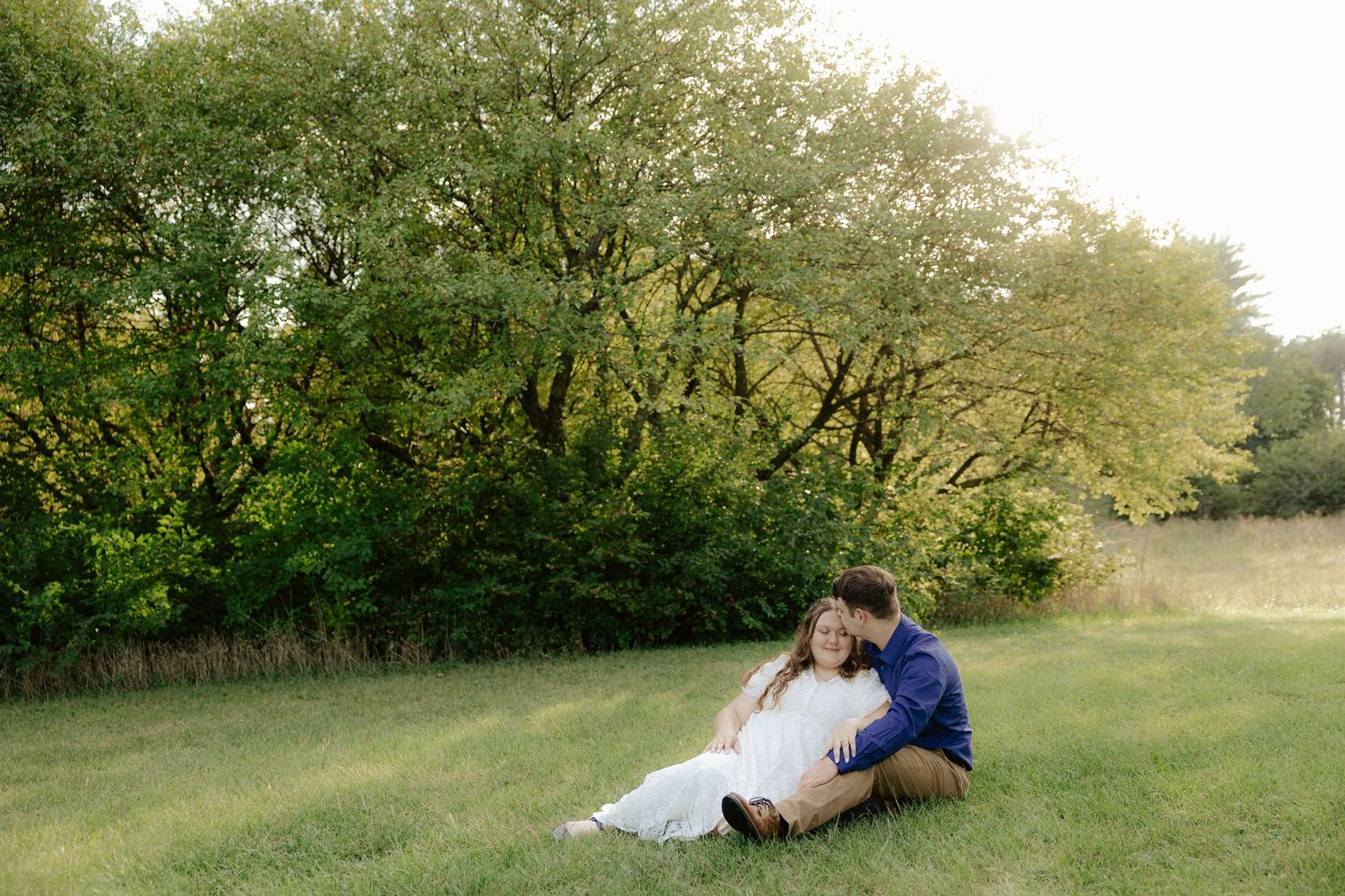 A couple sits together on grass in a natural outdoor setting surrounded by green trees and foliage at Pioneers Park in Lincoln Nebraska.