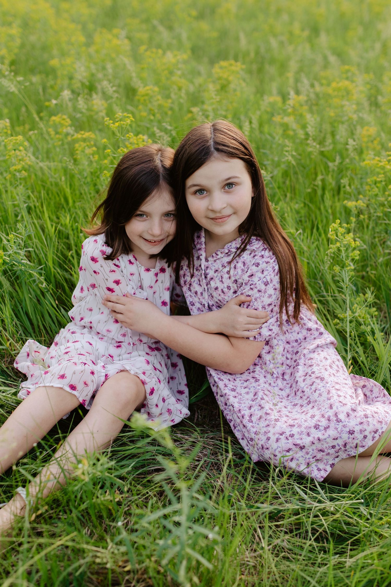 Two children in floral dresses sit together in a grassy meadow with wildflowers on a sunny day.