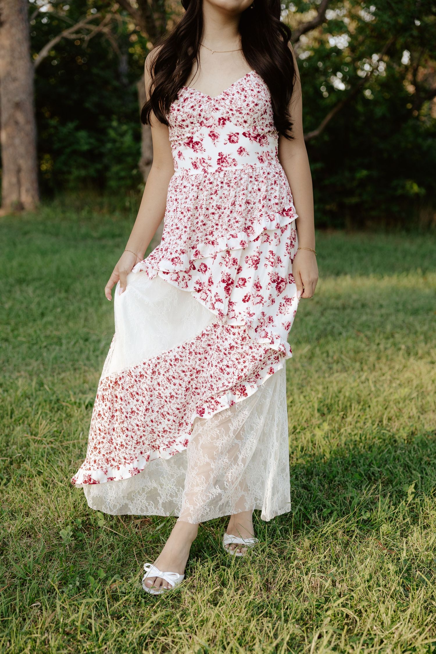 A girl in a white and red floral dress poses outdoors under blooming trees on a sunny day for her senior photos.