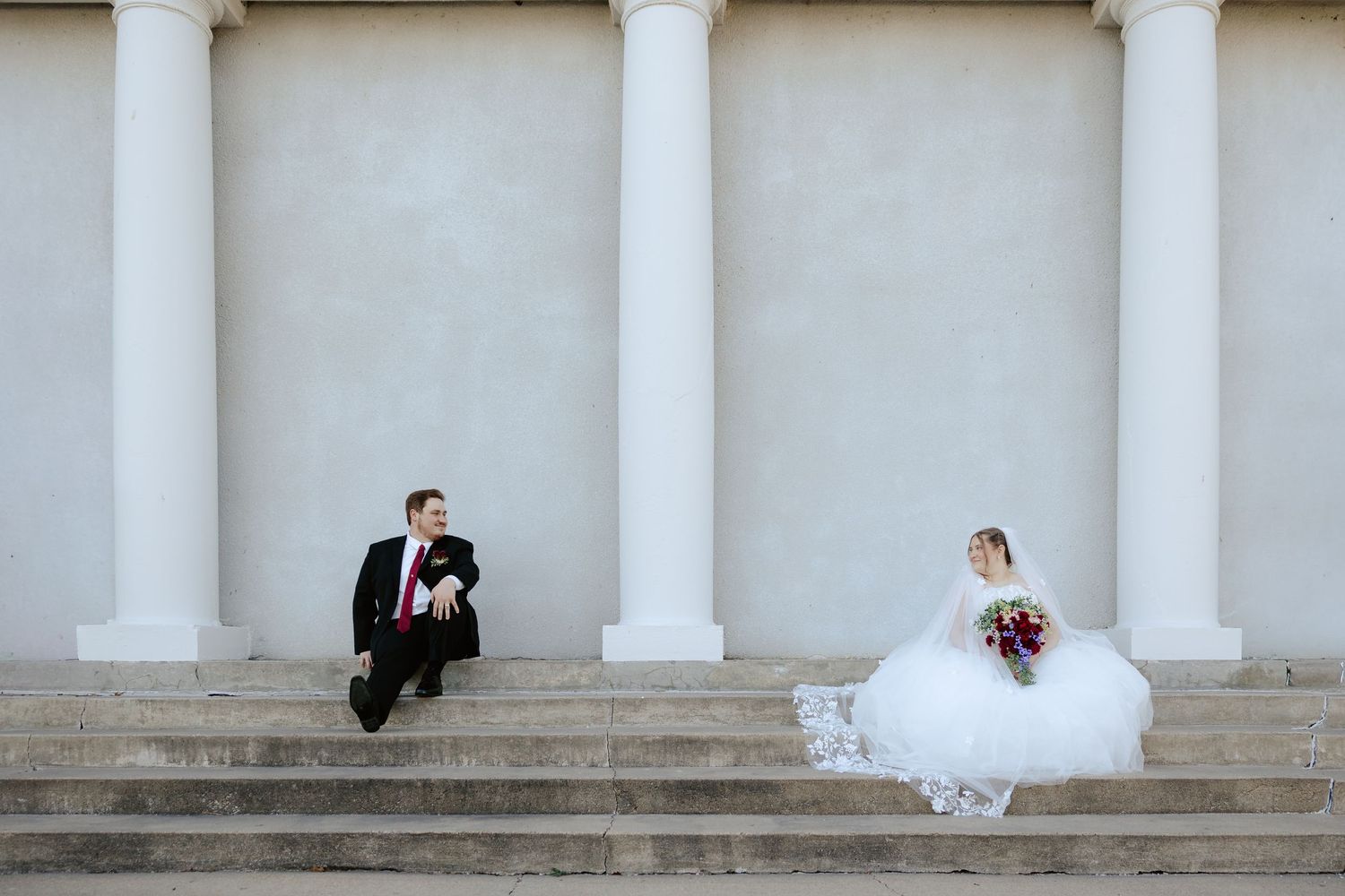 Bride and groom posing between white classical columns at the Auld Pavilion in Antelope Park in Lincoln Nebraska.