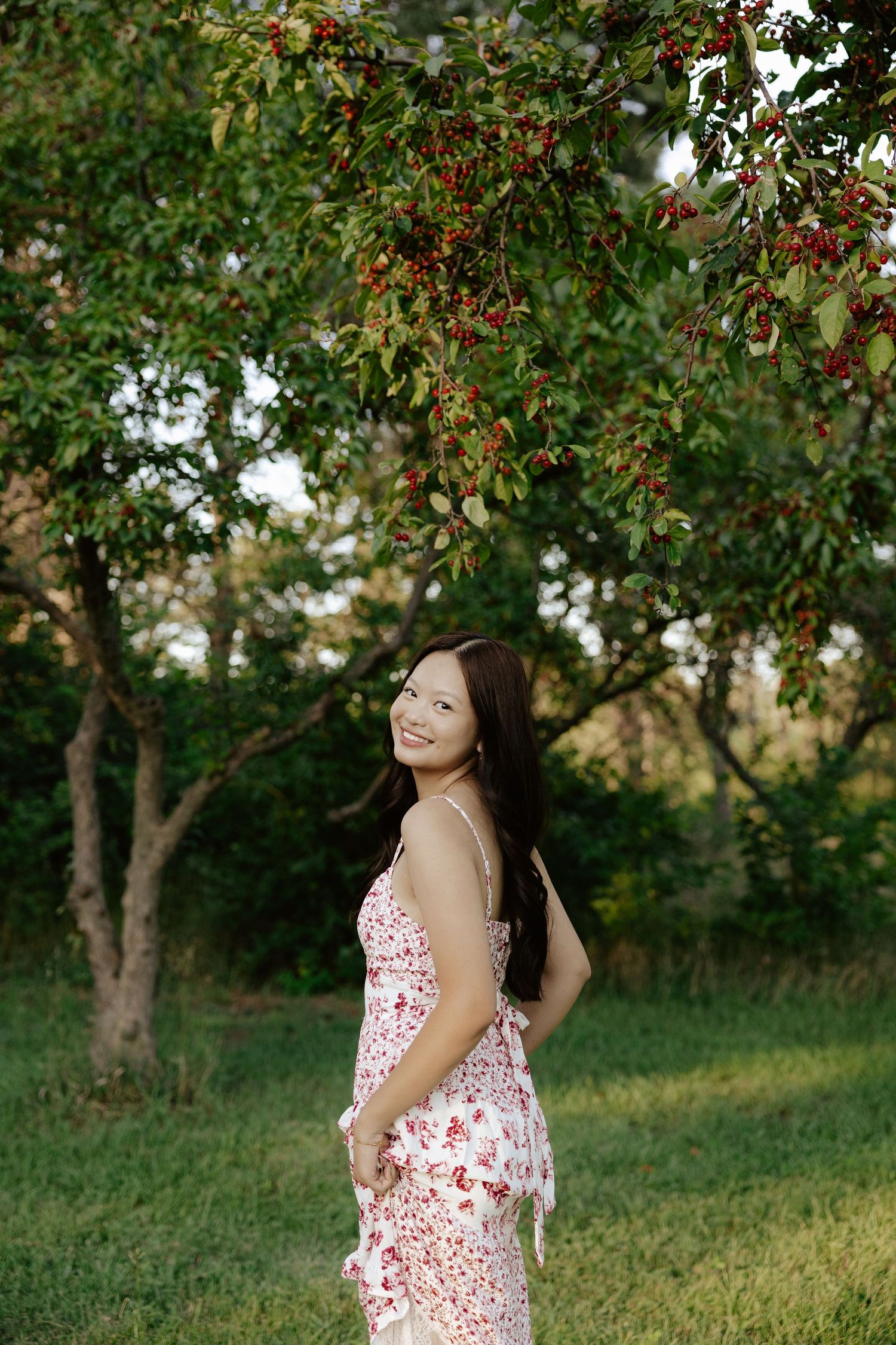 A girl in a white and red floral dress poses outdoors under blooming trees on a sunny day for her senior photos.