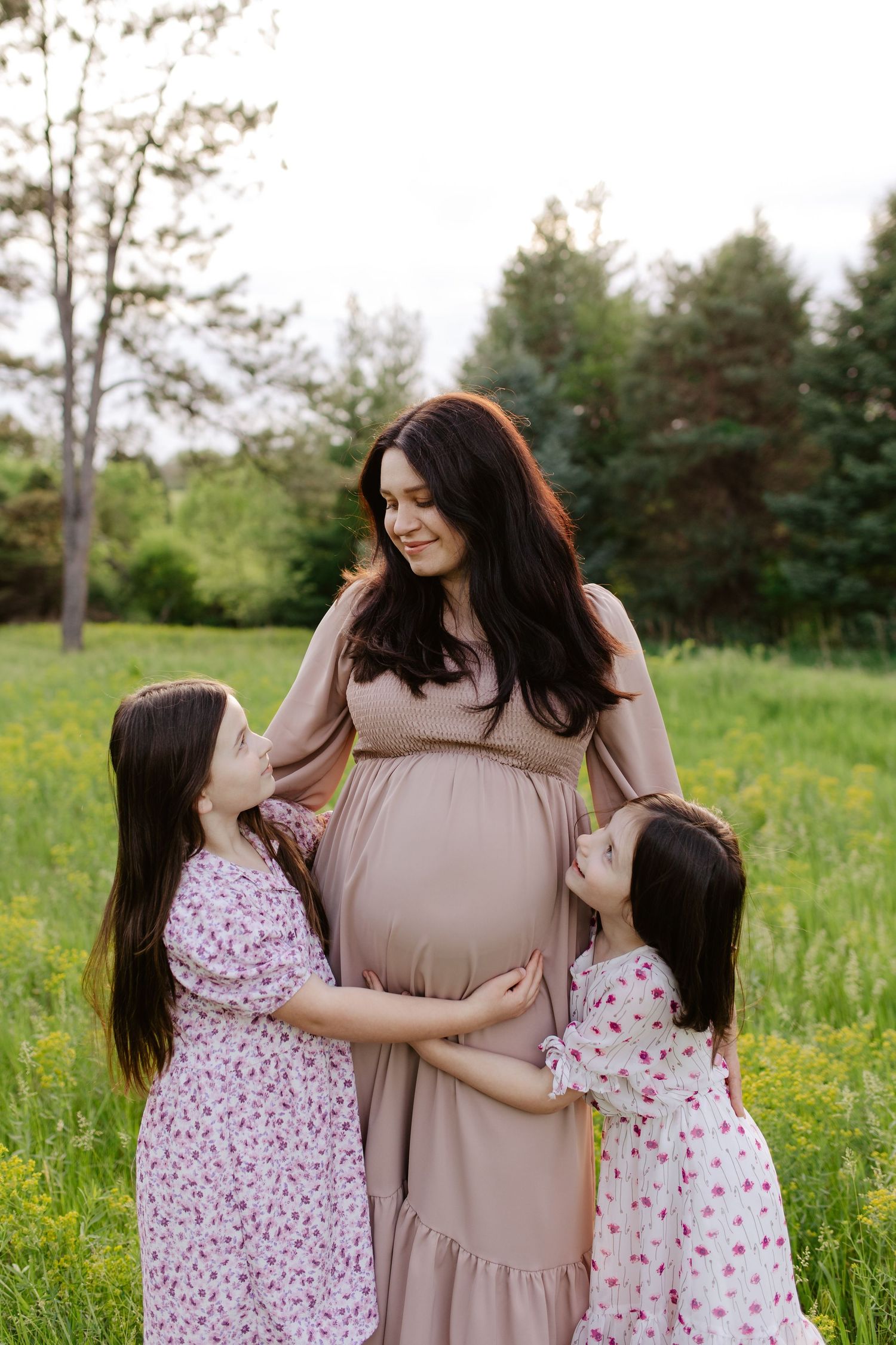 Mother and daughters share a tender moment in a grassy field with pine trees, as they embrace during a maternity photoshoot.